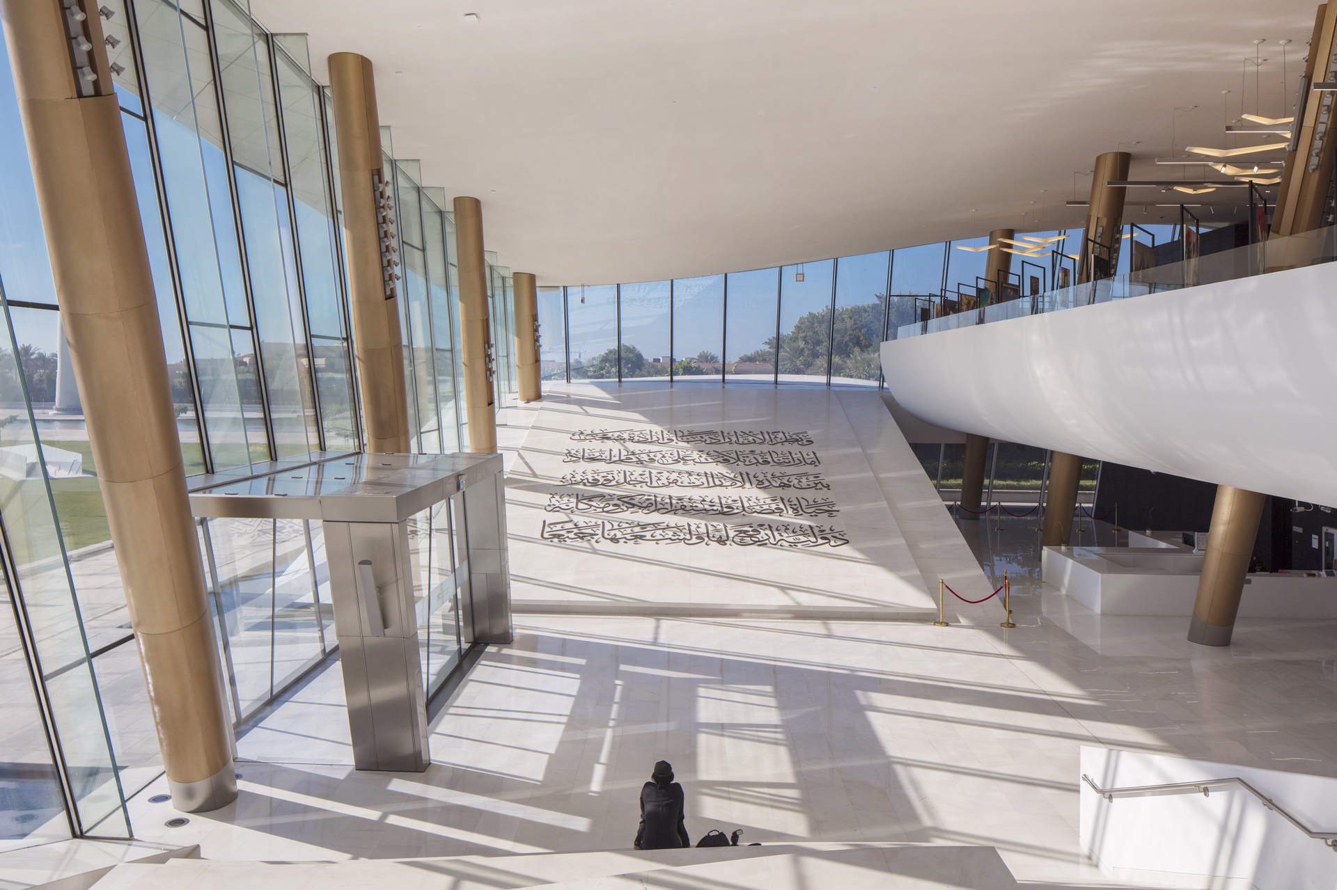 Spacious modern interior with large windows, golden pillars, Arabic calligraphy on white floor, and a person seated near stairs.