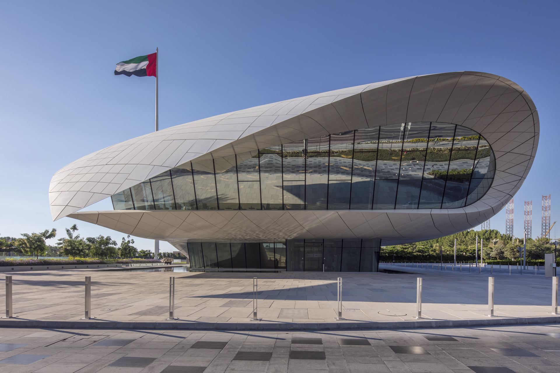 Modern futuristic building with curved white exterior and large glass windows under clear blue sky, UAE flag flying nearby.