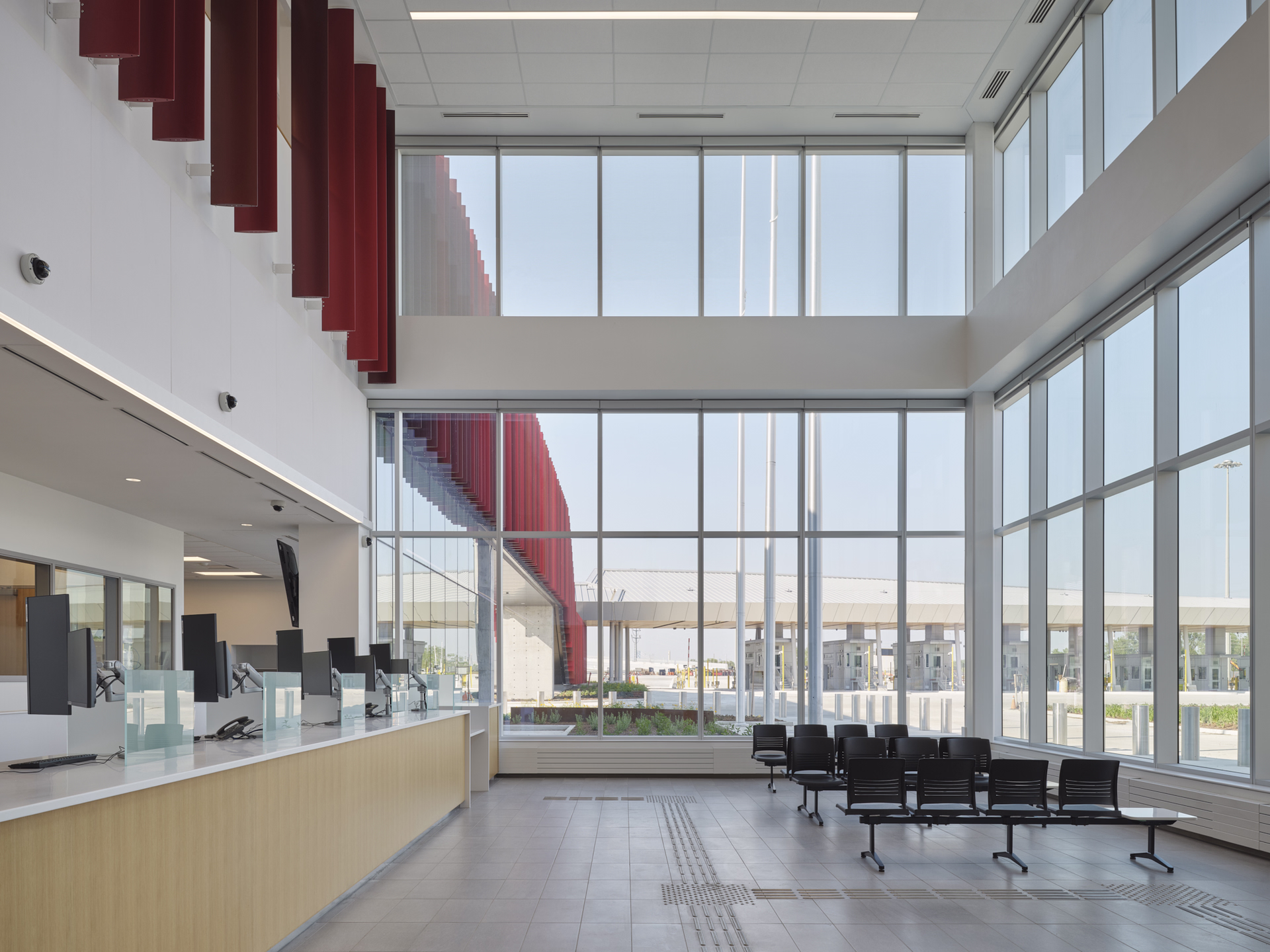 Modern, empty waiting area with large floor-to-ceiling windows, rows of black chairs, and a reception counter with multiple computer monitors.