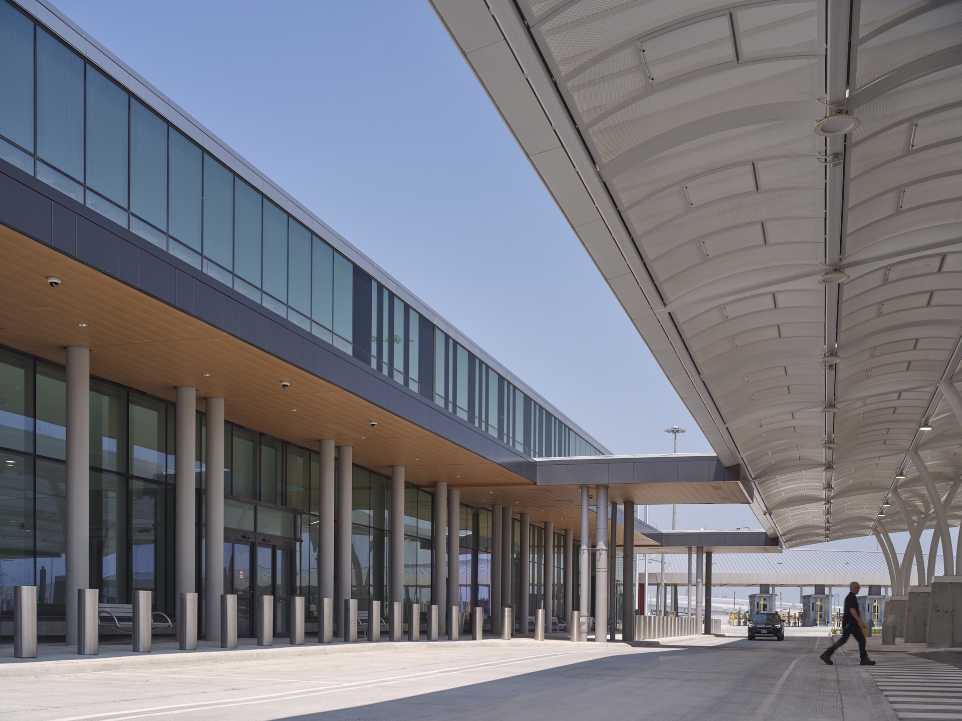 Modern airport terminal exterior with glass walls, metal columns, covered walkway, and a person crossing the road.