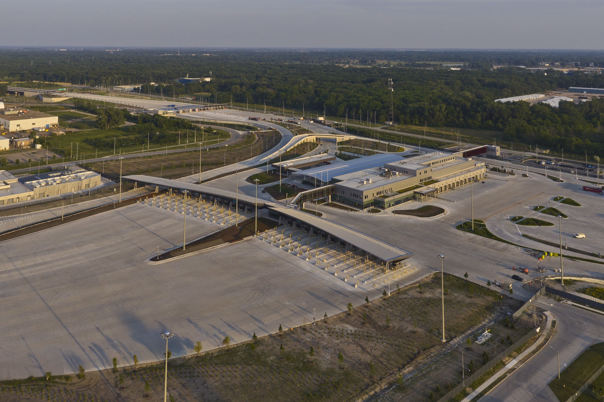 Aerial view of a large, empty toll plaza with multiple lanes and a modern building next to a highway surrounded by green forest.