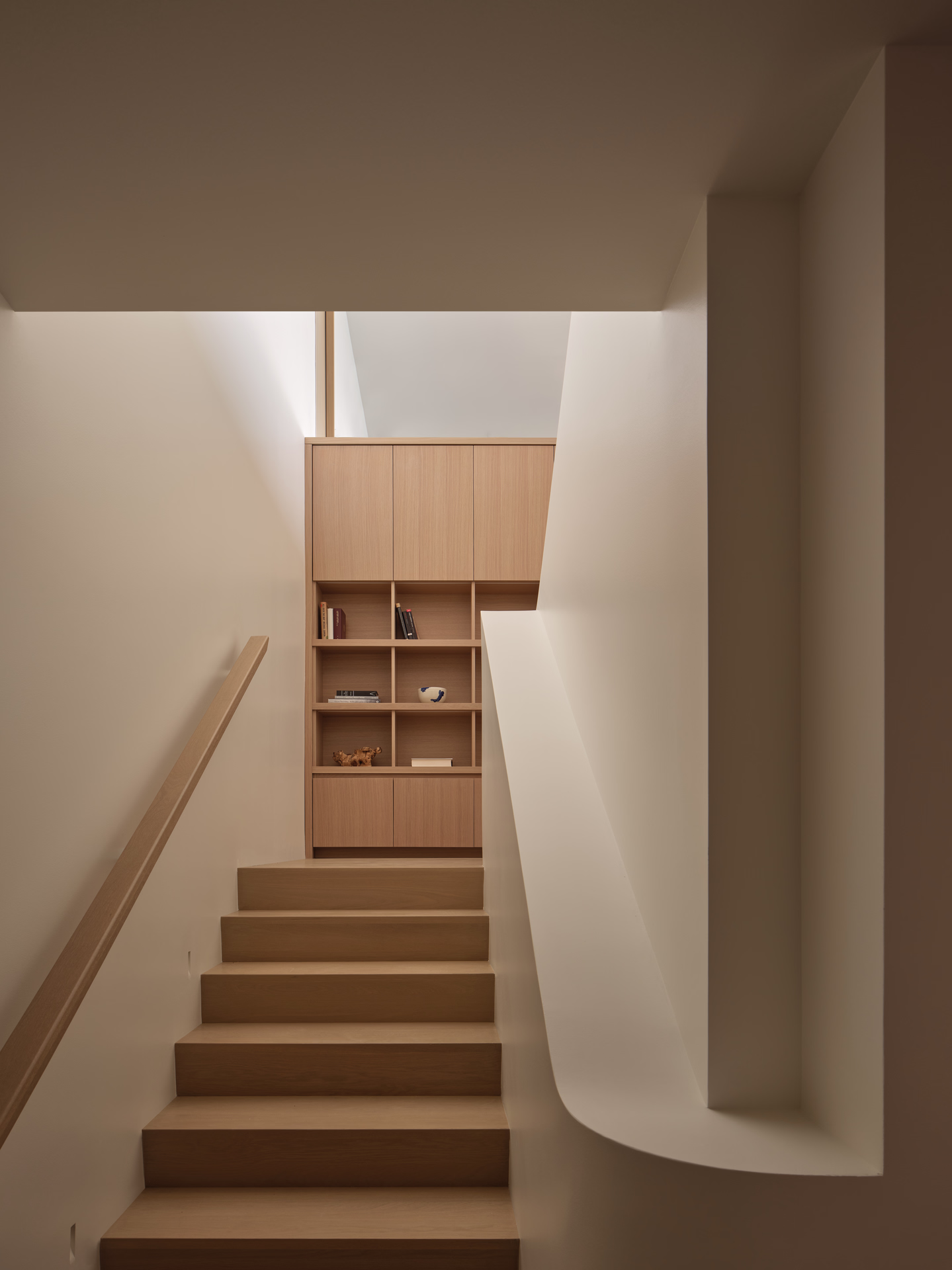 Minimalist wooden staircase leading up to a built-in wooden shelving unit with books and decorative items.