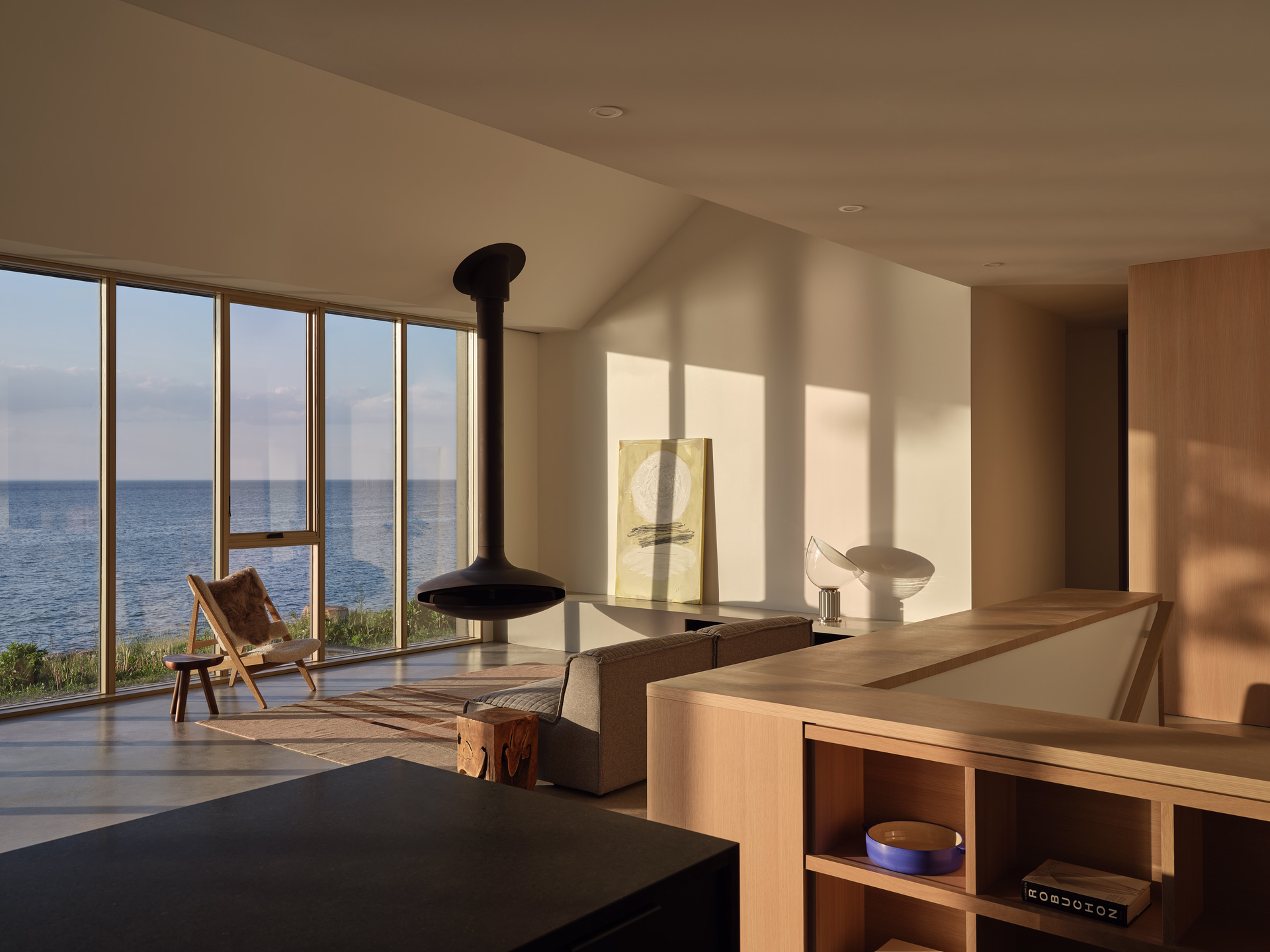 Modern living room with large floor-to-ceiling windows overlooking the ocean, a hanging black fireplace, a wooden chair with fur, and minimalist decor.