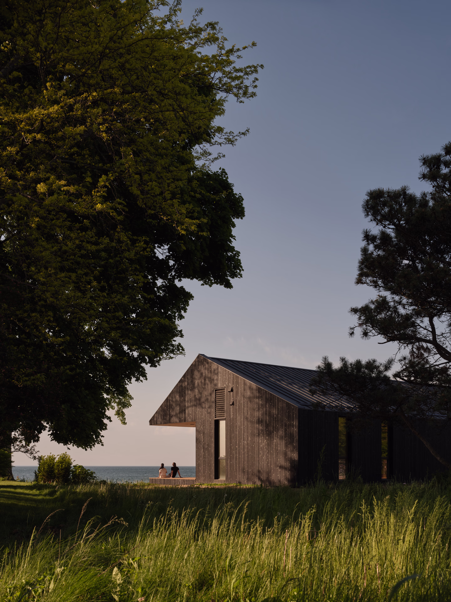 Modern dark wooden house near a grassy area with two people sitting on its deck overlooking a calm sea at sunset.