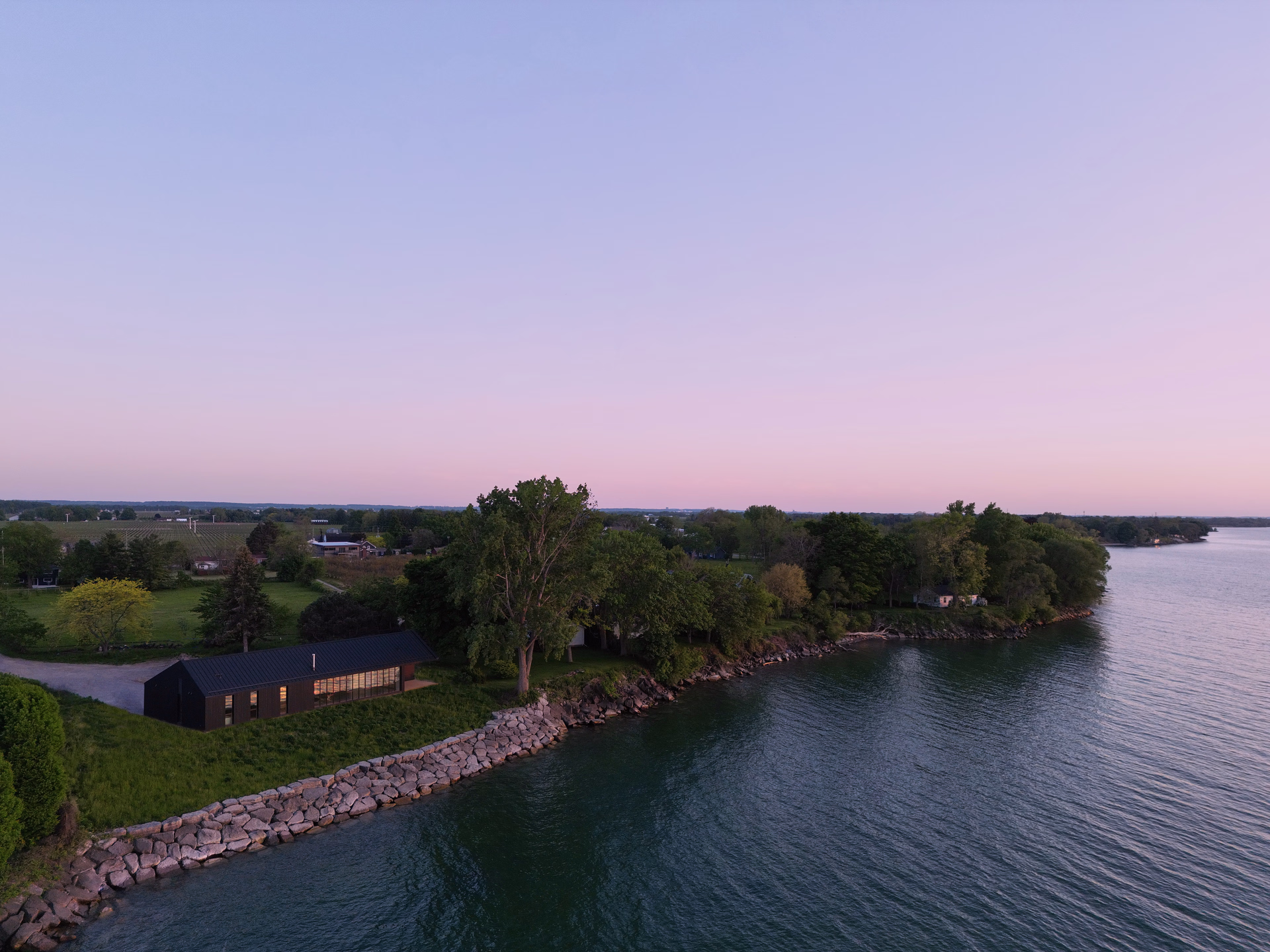 A modern black house situated near a rocky shoreline with trees and calm water at sunset.
