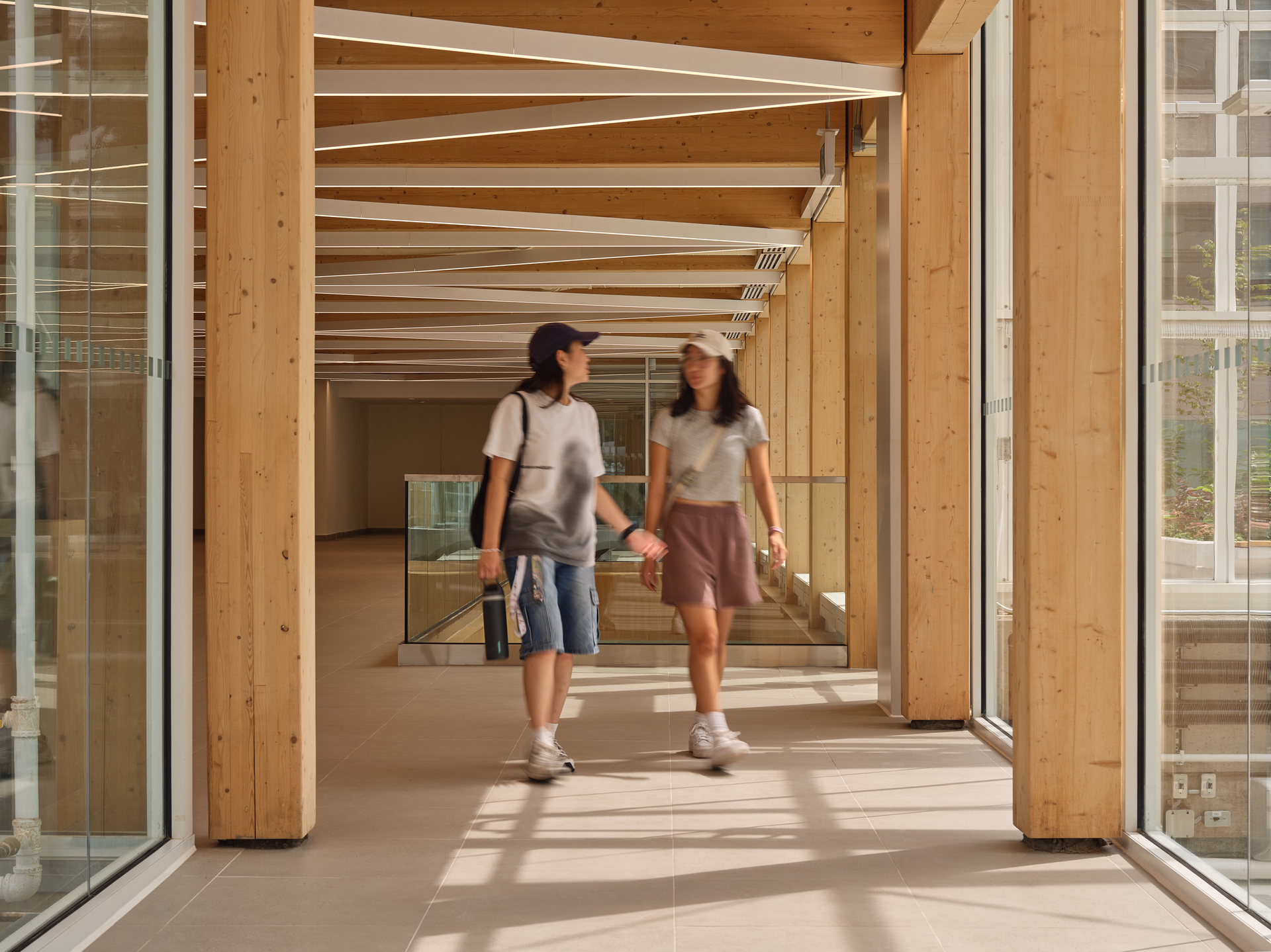 Two young women walking and talking in a bright corridor with wooden pillars and large windows.