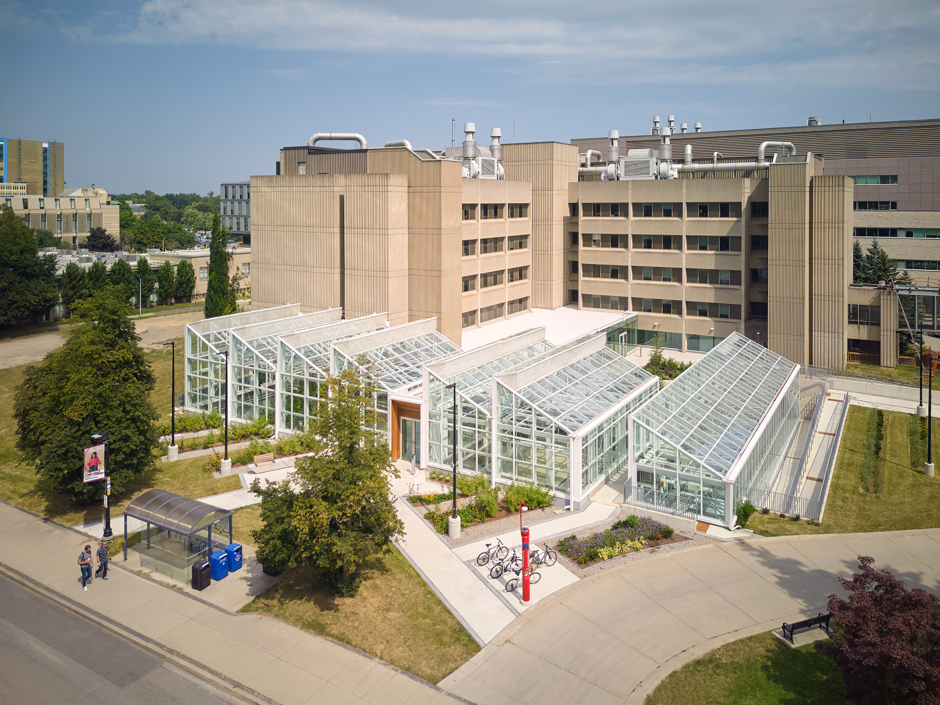 Aerial view of a modern glass greenhouse building adjacent to a beige multi-story institutional building, surrounded by walkways and greenery.