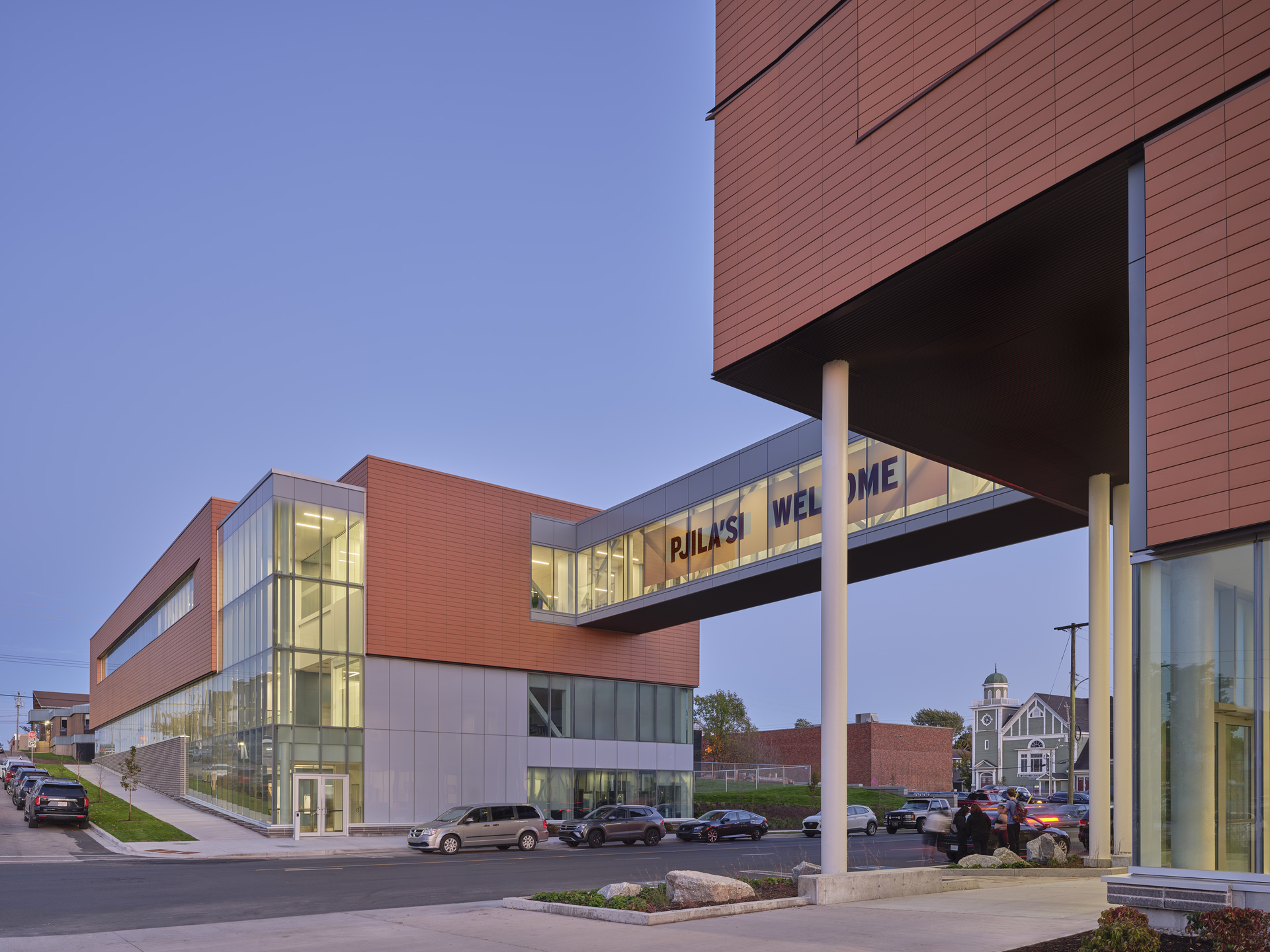 Modern school building with a glass walkway displaying the text 'Pjila'si Welcome' over a street with parked cars at dusk.