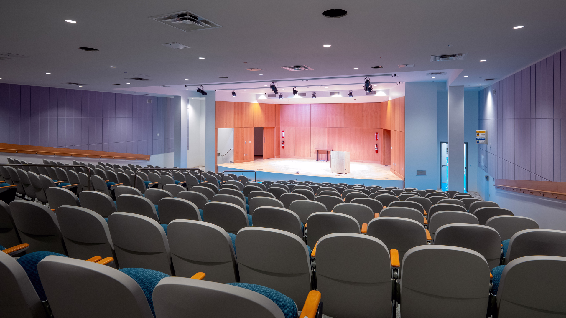 Empty auditorium with rows of gray seats all facing a well-lit wooden stage with a podium and a table.
