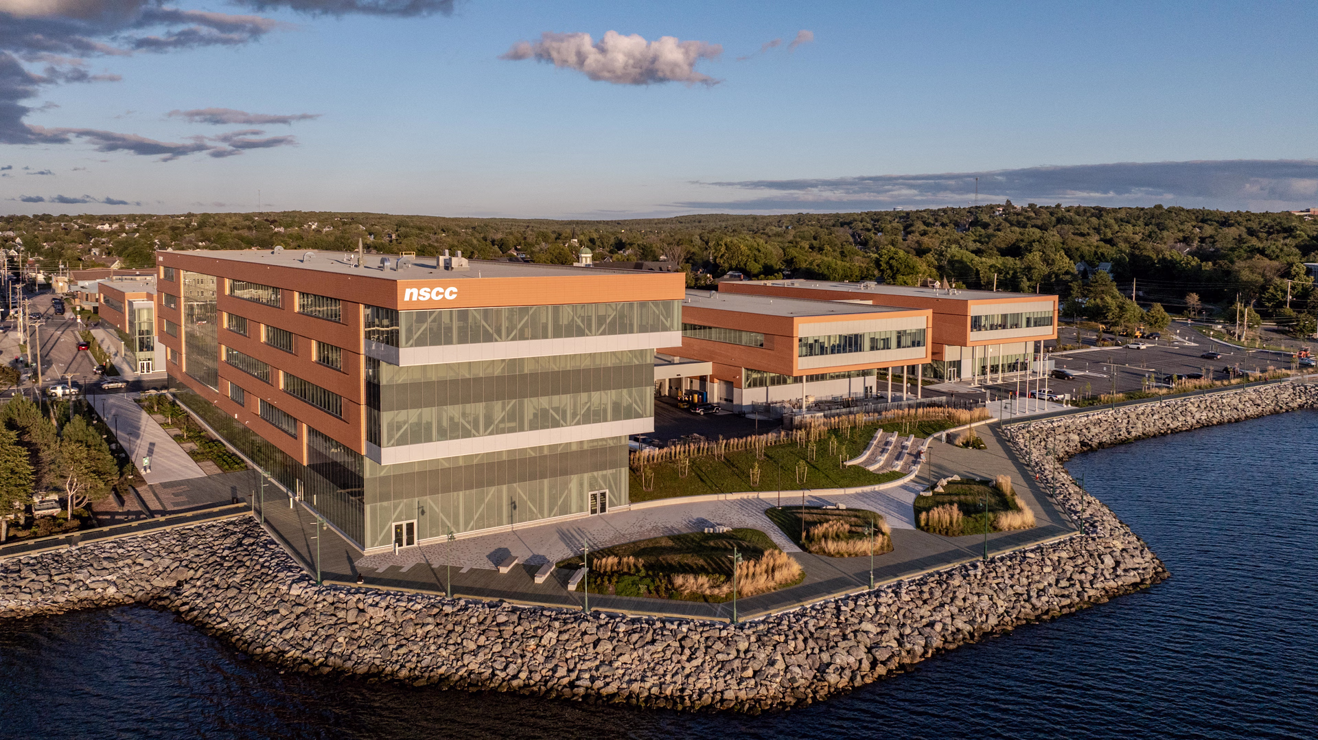 A modern waterfront building complex with NSCC logo, surrounded by a stone embankment and adjacent greenery under a partly cloudy sky.