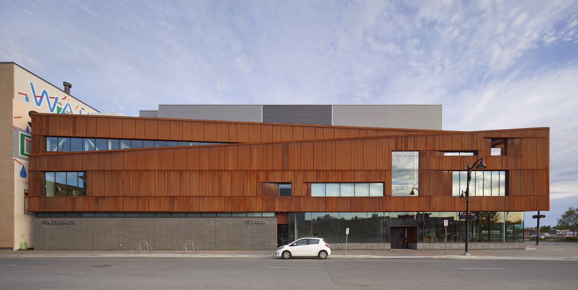 Modern building with rust-colored metal panels, large irregular windows, gray stone base, and a white car parked in front on the street.