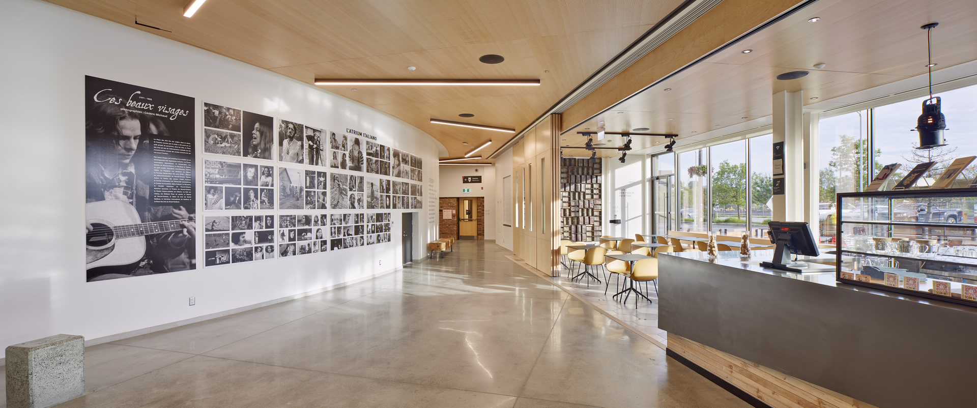 Modern interior hallway with a photo gallery on the left wall and a café seating area with yellow chairs and large windows on the right.