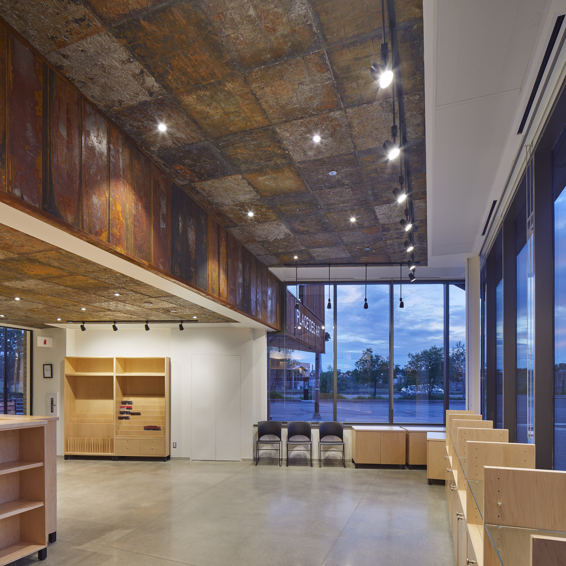 Modern interior with rusted metal ceiling panels, wooden shelves, three black chairs, and large windows showing an outdoor view.