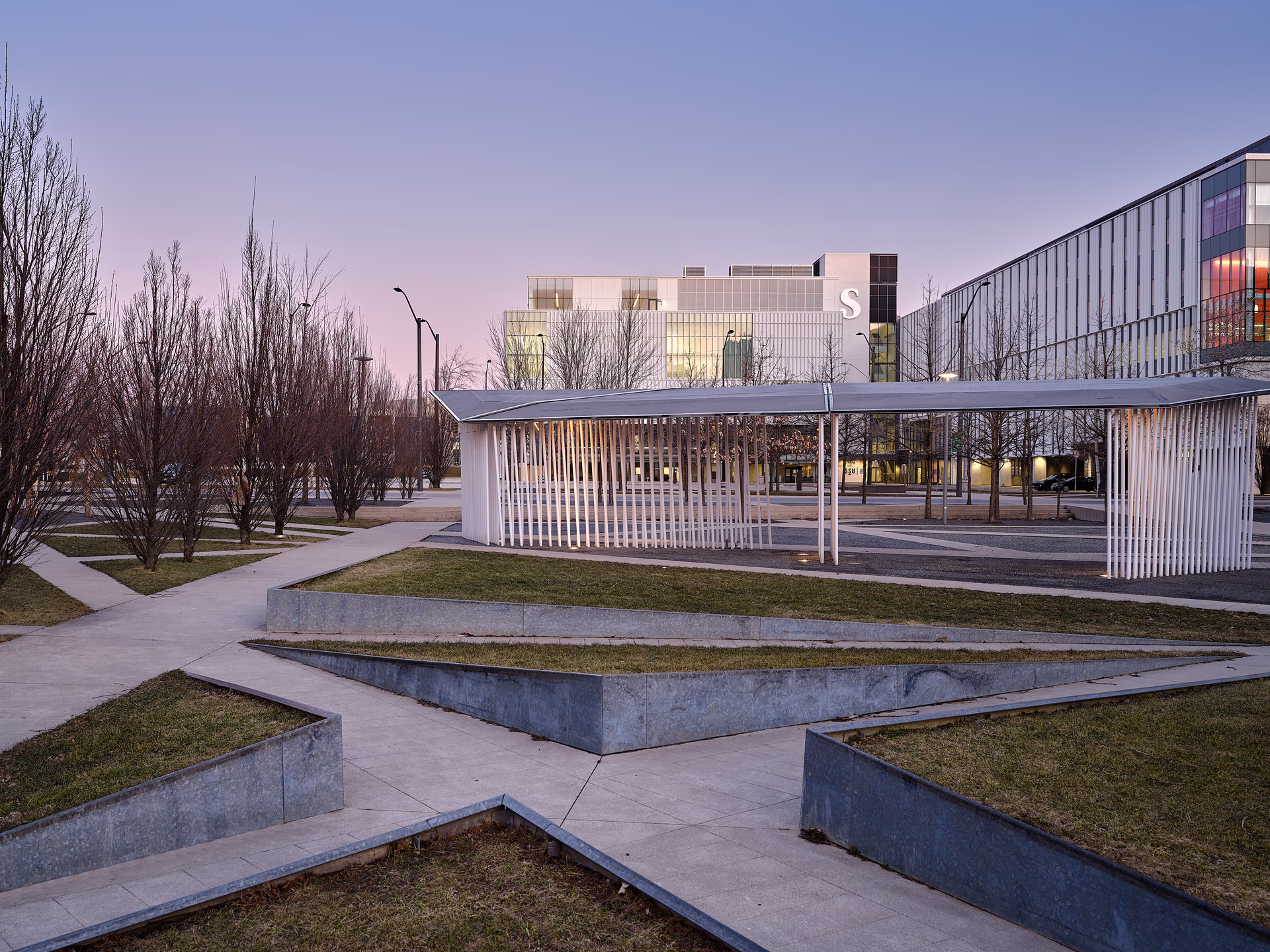 Modern urban park with geometric concrete paths, bare trees, and a white minimalist pavilion in front of glass-front buildings during twilight.