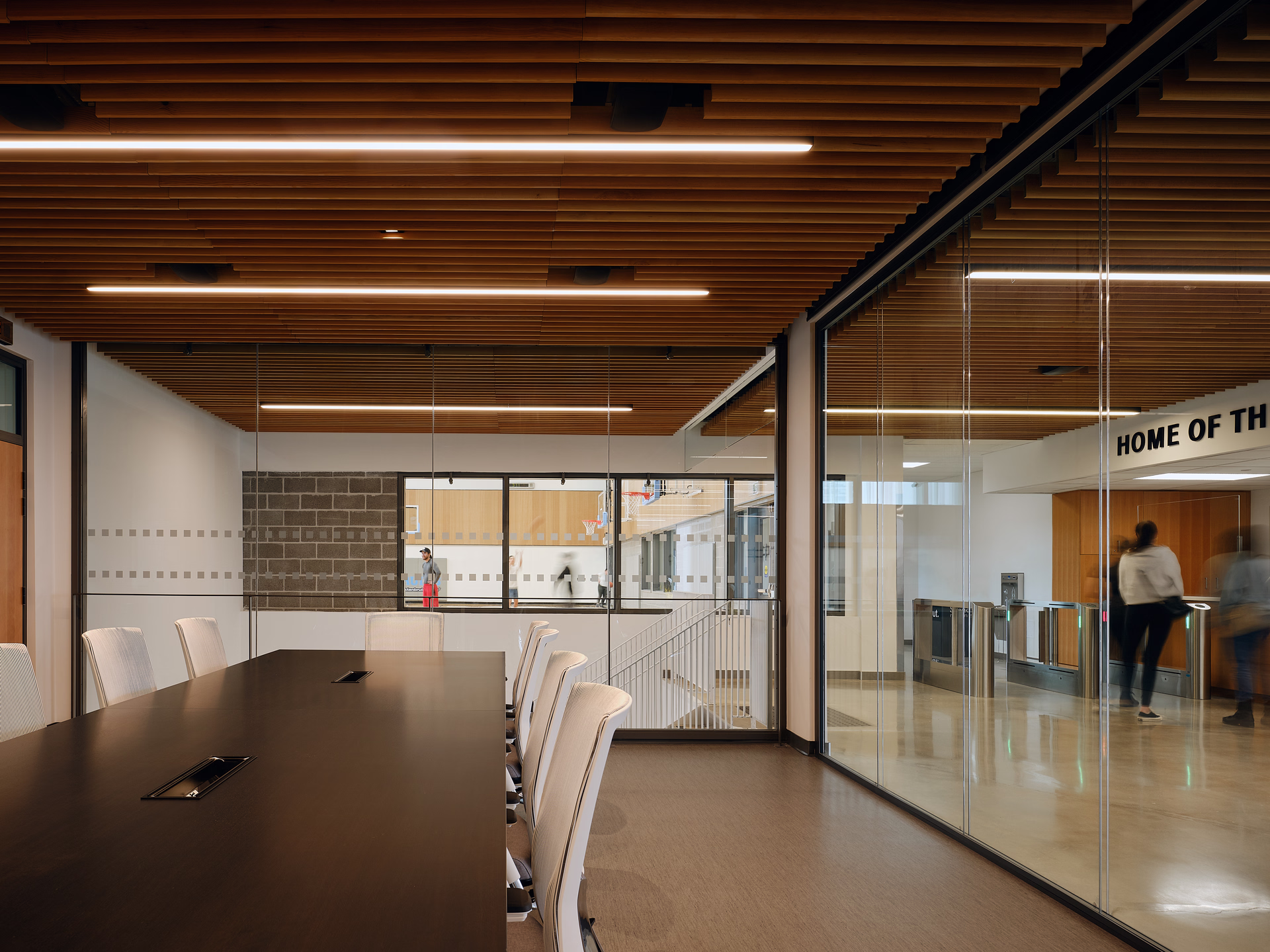 Modern conference room with long black table, white chairs, wooden slatted ceiling, and glass walls overlooking a hallway and basketball court.