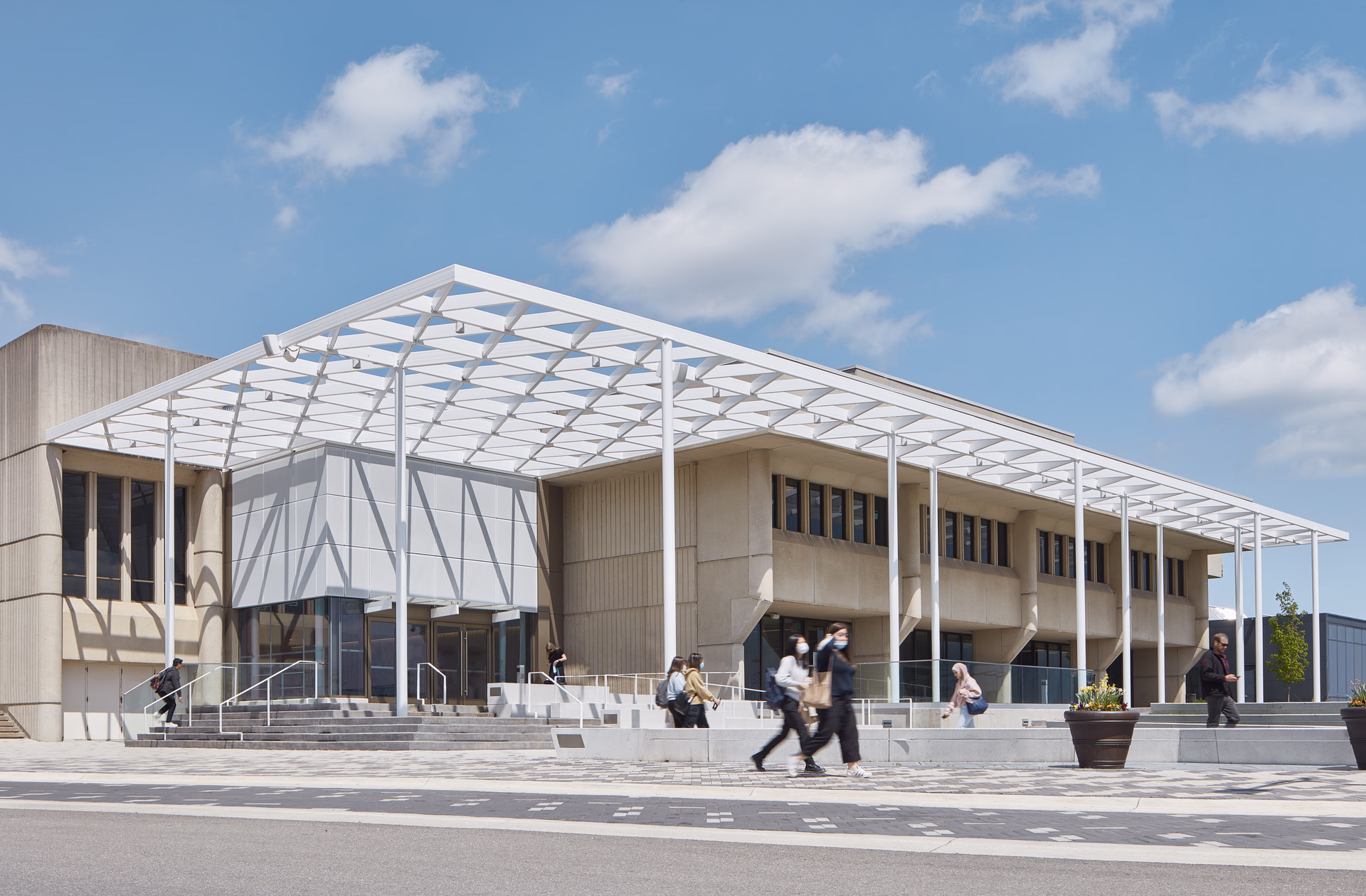 Modern concrete building with a white metal canopy and people walking and sitting around the entrance on a sunny day.