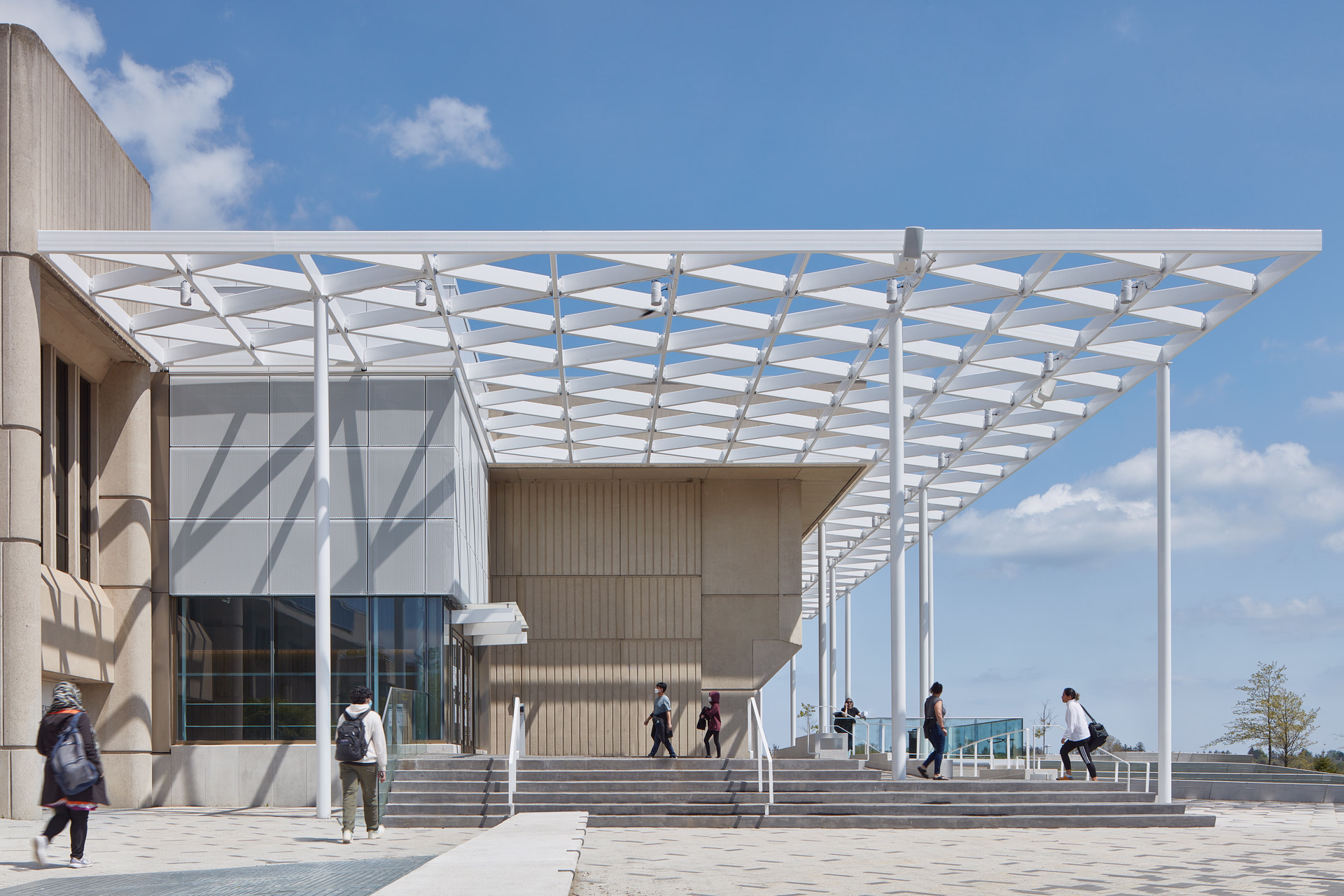 People walking near a modern building entrance with a white geometric canopy structure under a blue sky.