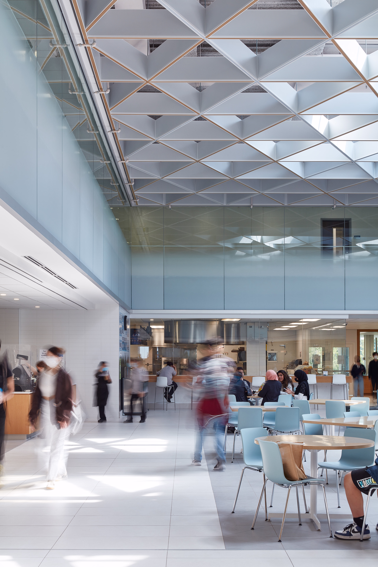Modern cafeteria with glass walls, white tiled floor, blue chairs, and blurred people walking and sitting.