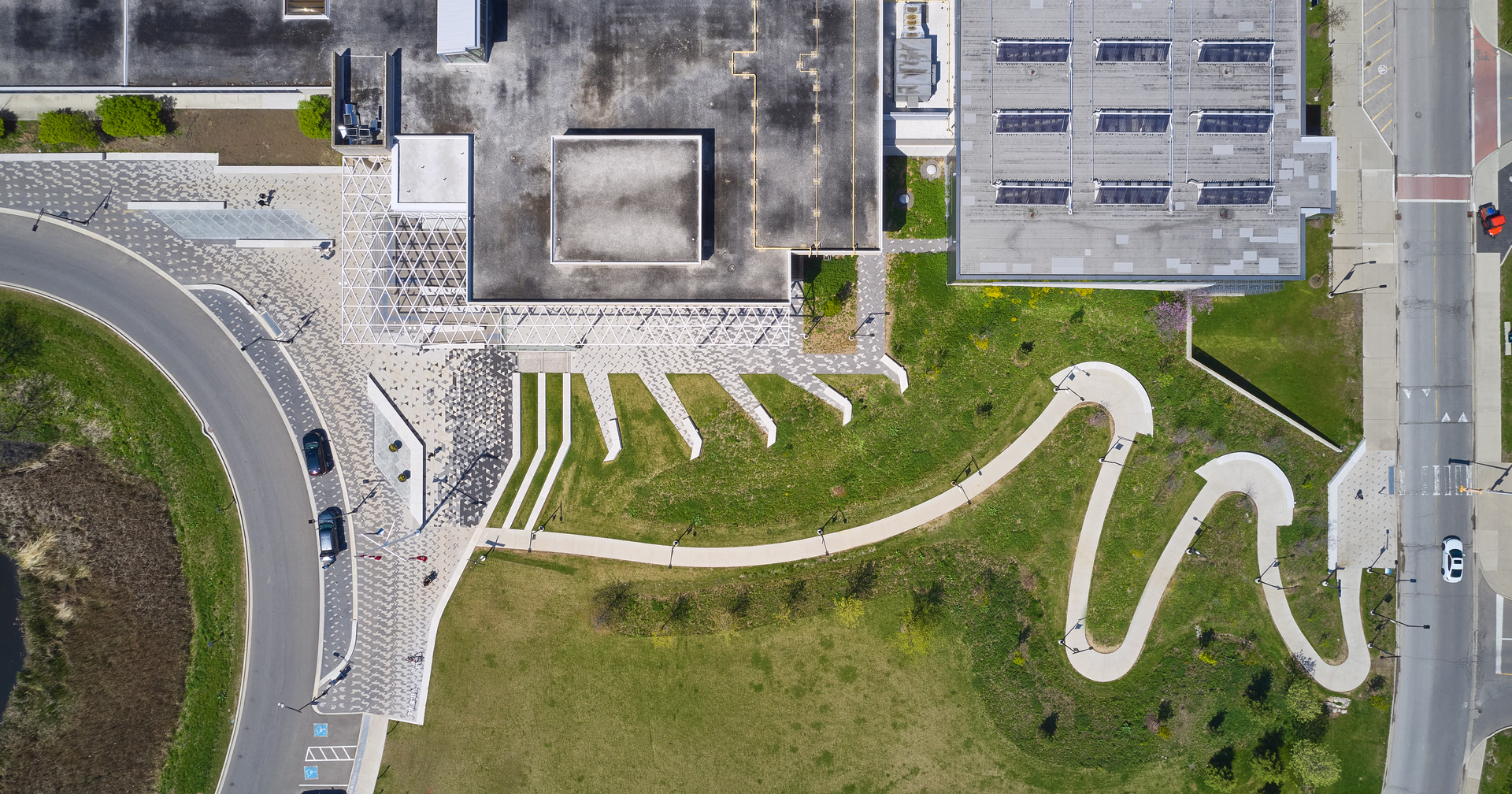 Aerial view of a building complex with rooftop structures, paved walkways, curved road, and green lawns with scattered trees and bushes.
