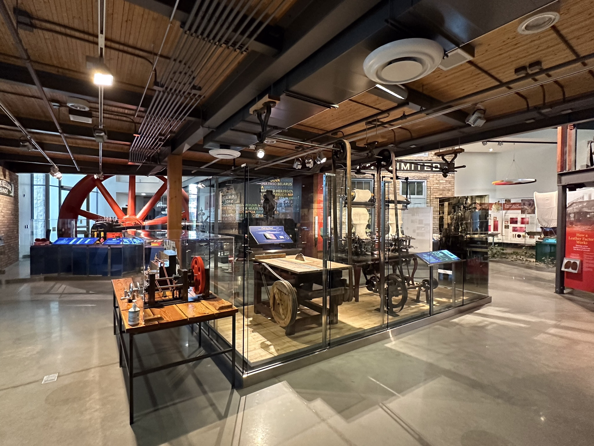 Industrial museum exhibit featuring vintage machinery and a large red flywheel in a spacious, well-lit room with polished concrete floors.