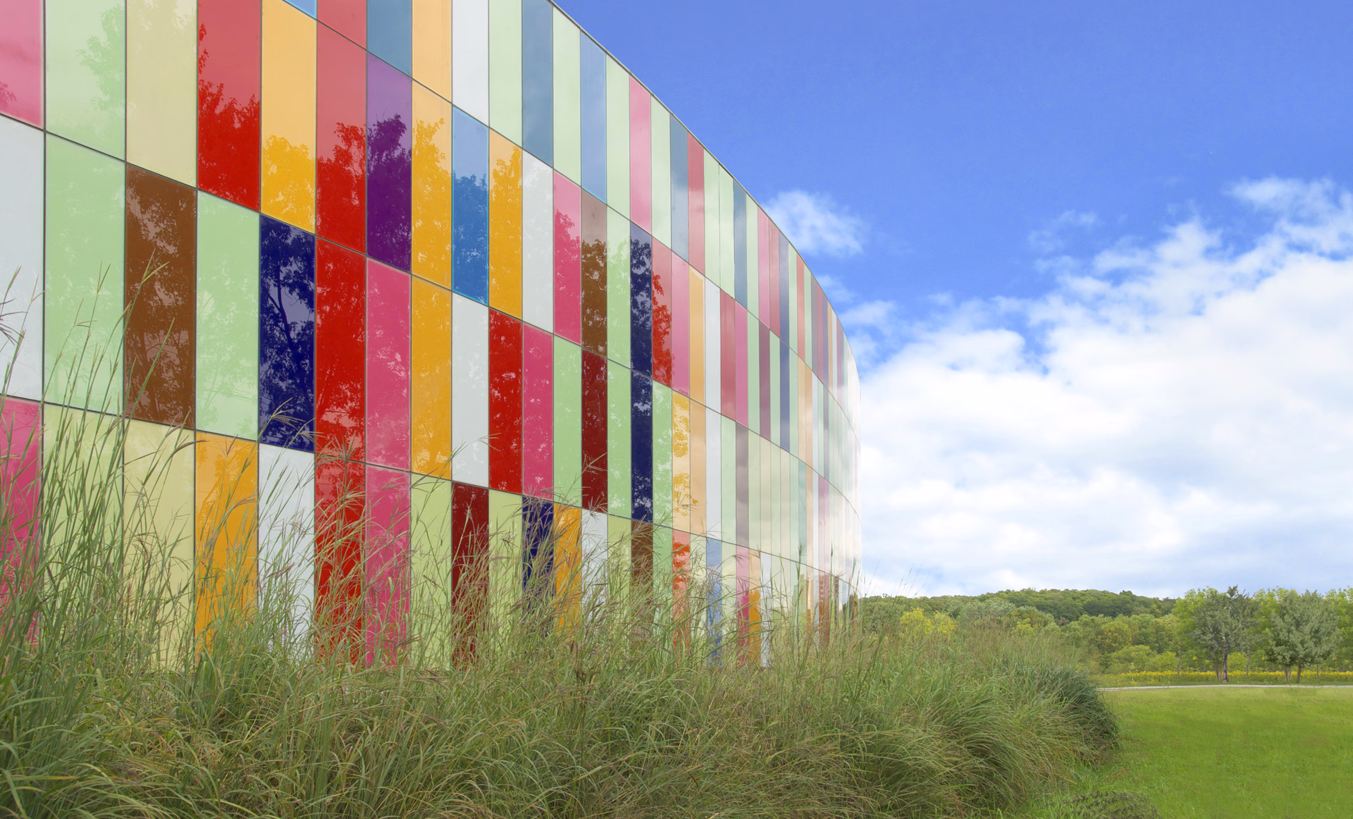 Curved building facade covered with colorful rectangular glass panels reflecting trees, with tall grass in the foreground and a blue sky with clouds above.