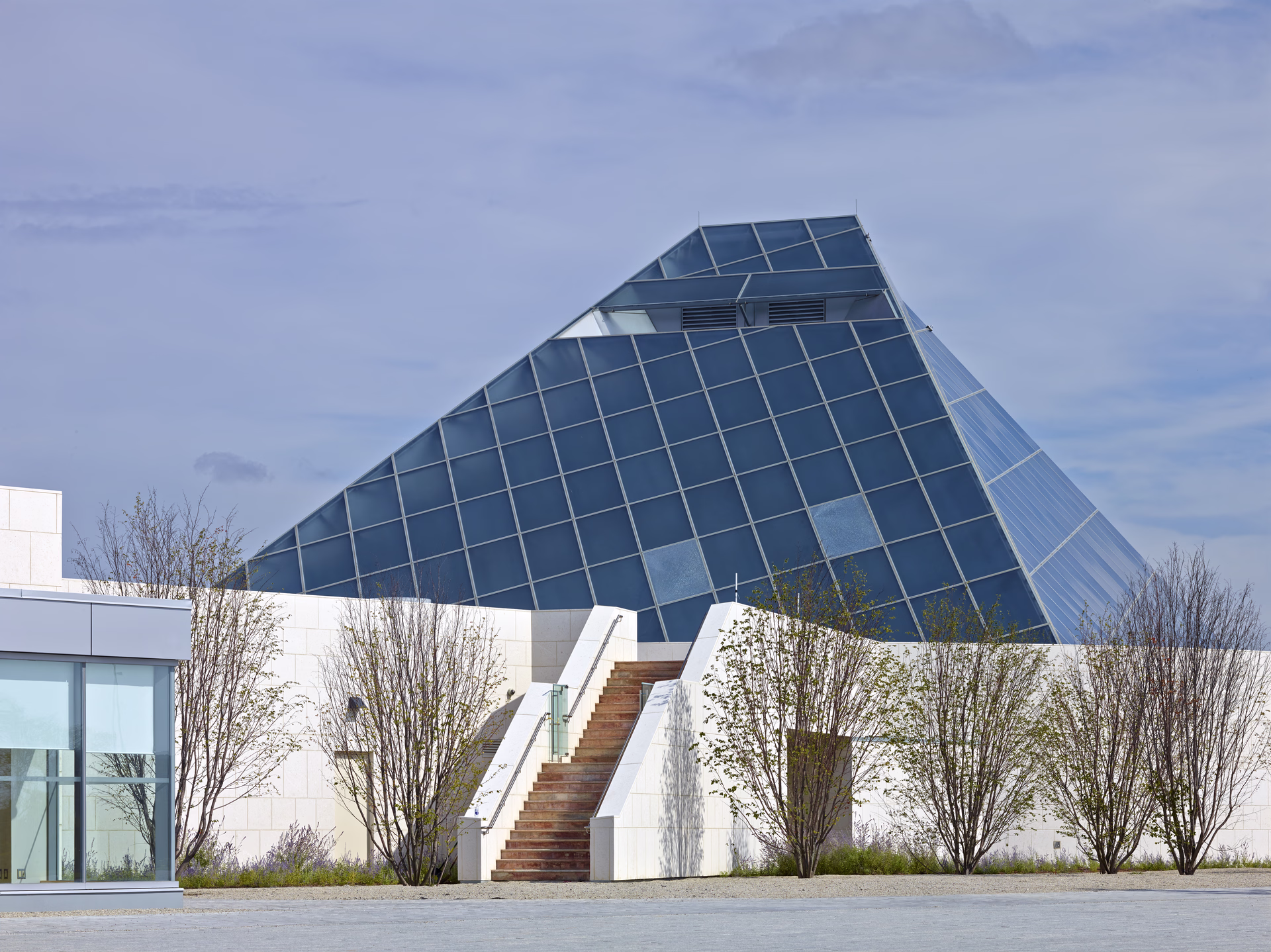 Modern building with a large, slanted glass pyramid roof and white walls, surrounded by young trees.