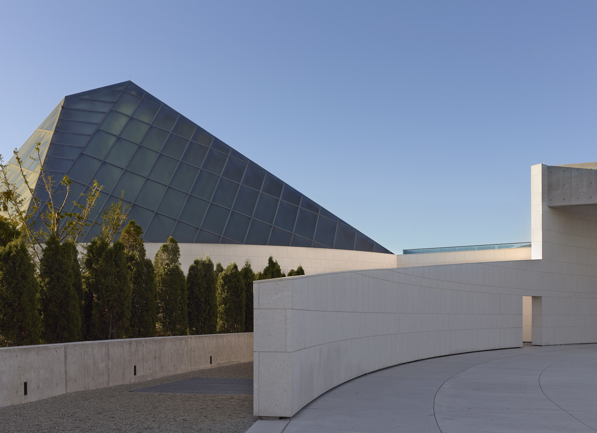 Modern architectural scene with a slanted glass building, curved white concrete walls, green trees, and clear blue sky.