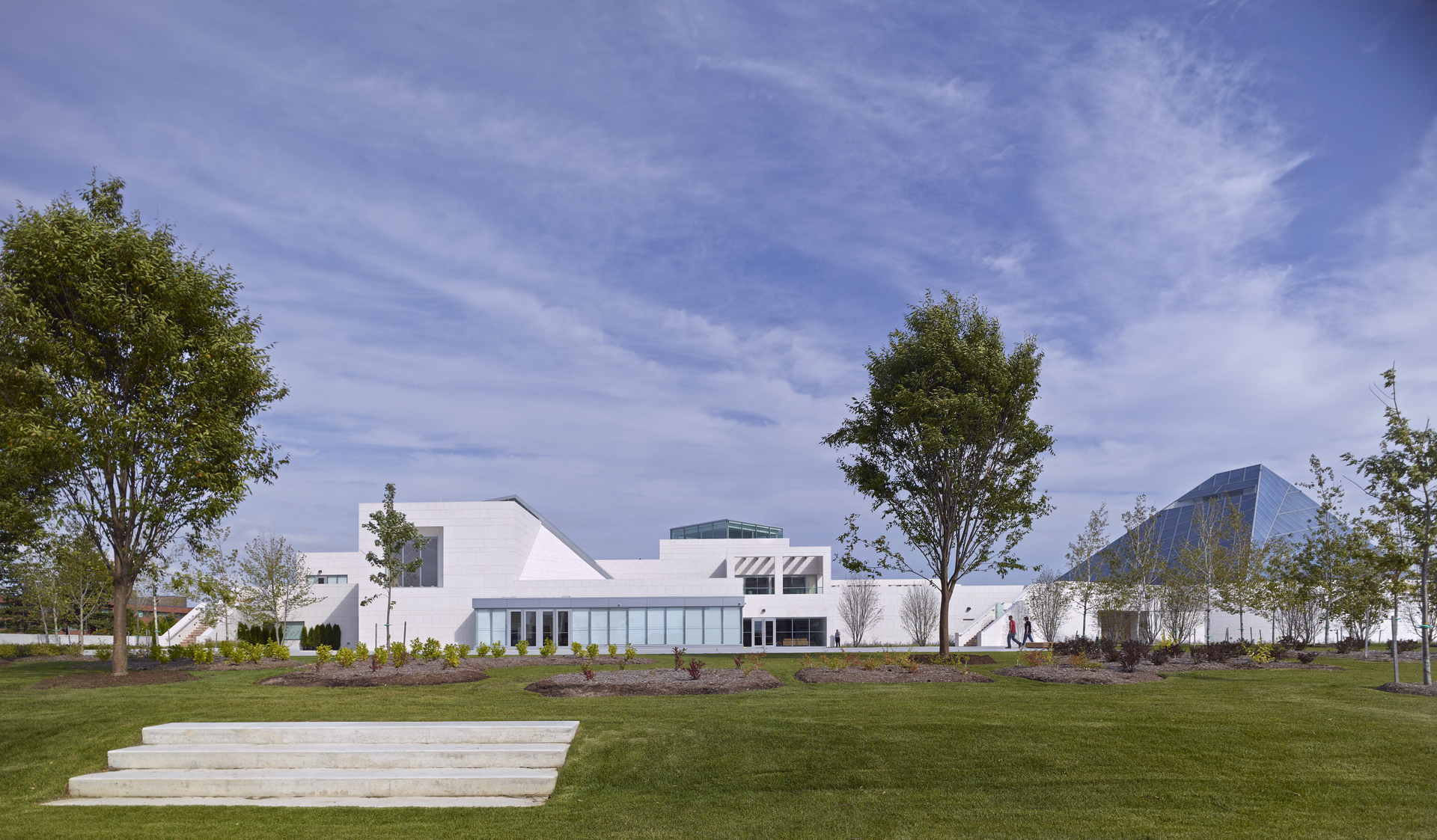 Modern white building complex with glass pyramid structure, surrounded by trees and green lawn under a partly cloudy blue sky.