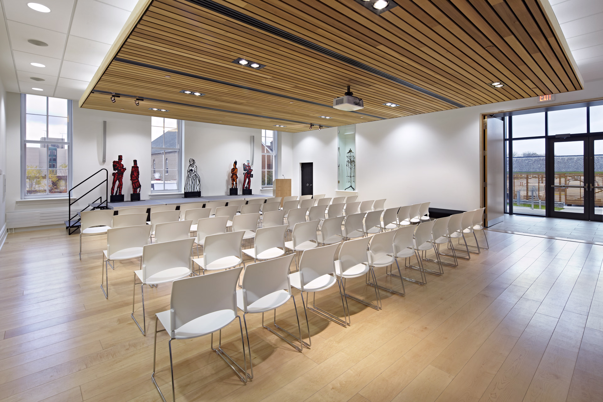 Modern conference room with rows of white chairs, a small stage with sculptures, wooden ceiling panels, and large windows.