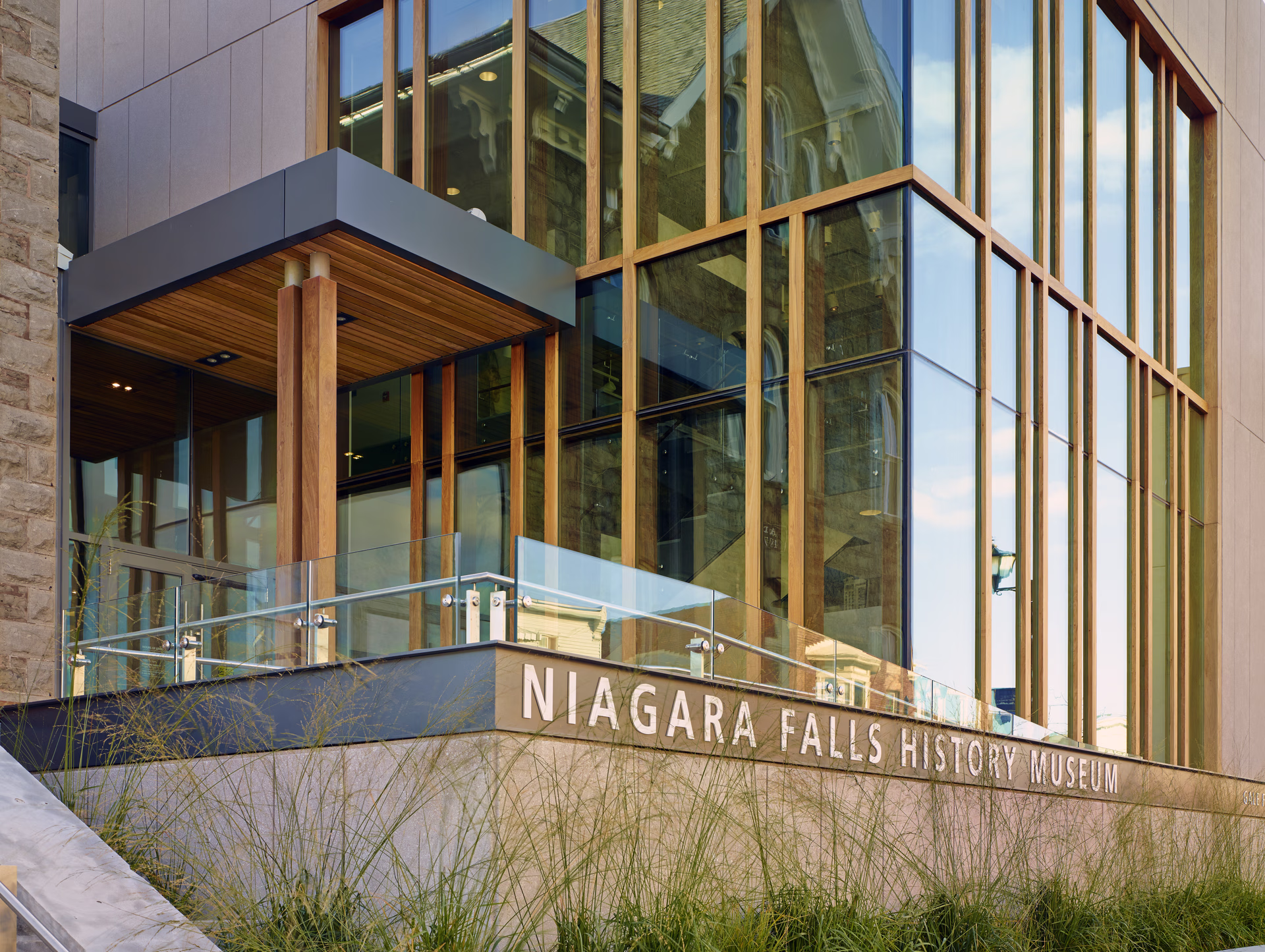 Modern glass and wood entrance of the Niagara Falls History Museum with reflections of older buildings in the windows.