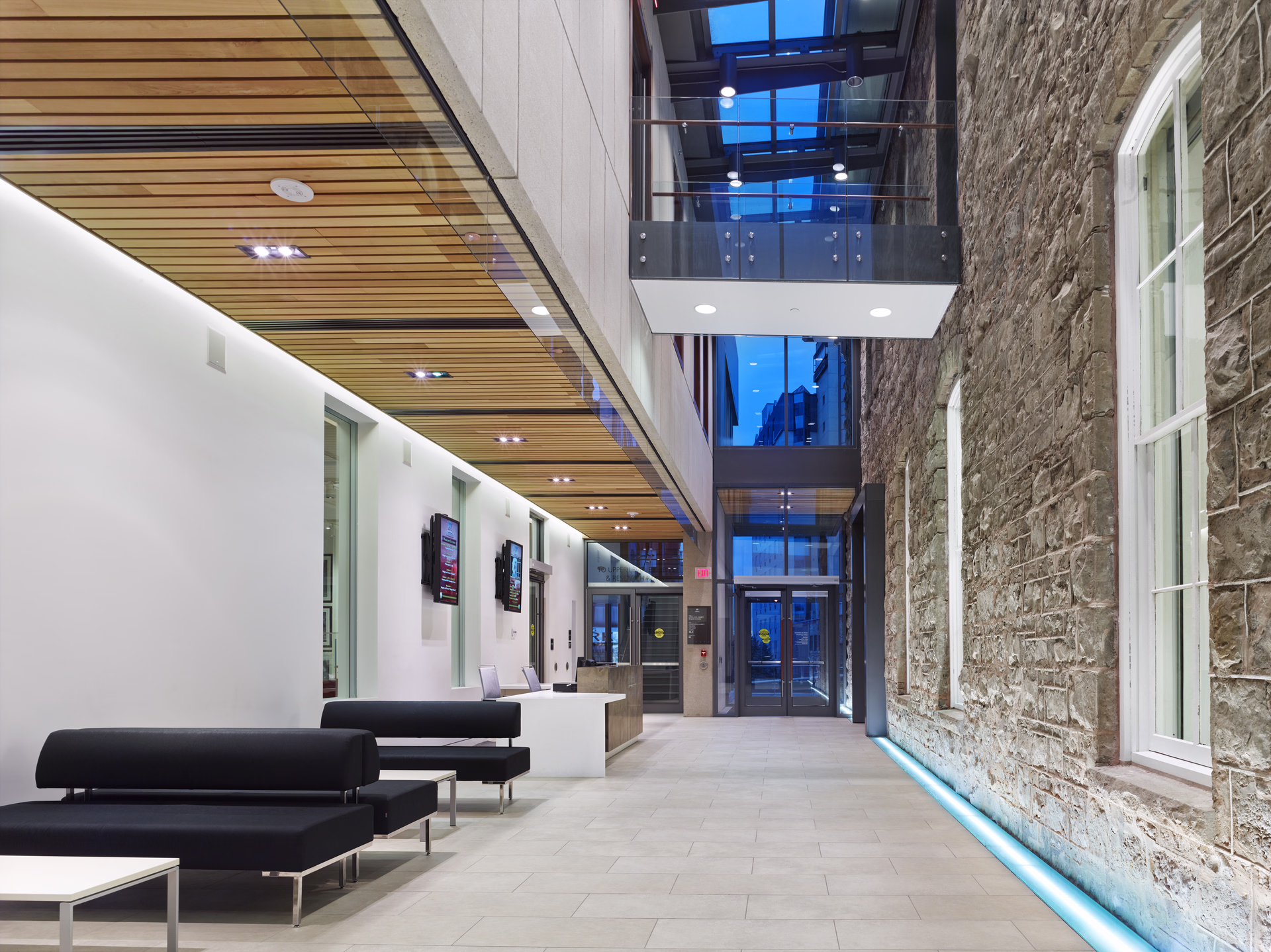 Modern lobby with black bench seating, light tiled floor, wood-paneled ceiling, and stone wall with white-framed windows.