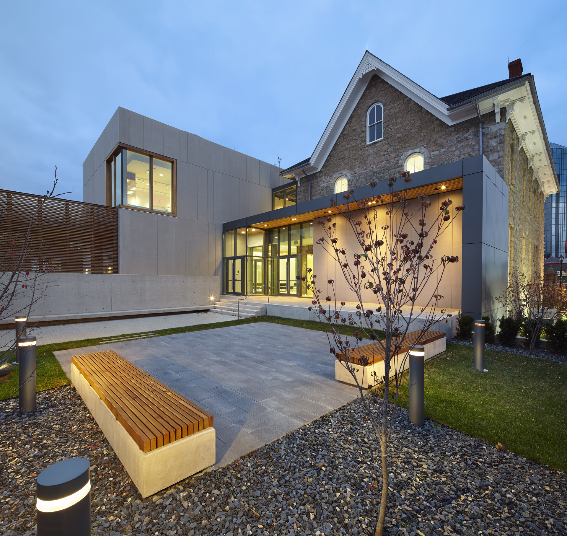 Modern building with large windows and stone facade illuminated at dusk, featuring wooden benches and landscaped courtyard.