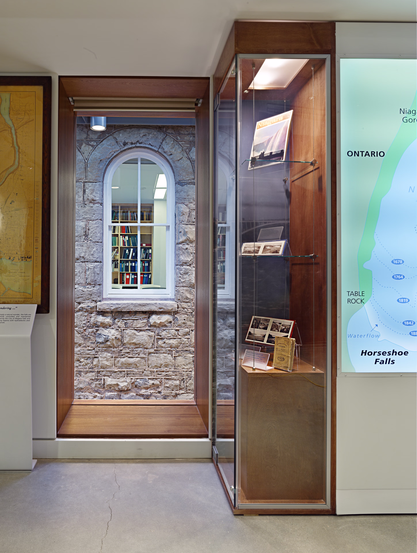 Museum display with a stone wall section featuring an arched window showing bookshelves inside, adjacent to a glass case holding historic photos and documents, and a nearby map highlighting Horseshoe Falls in Ontario.