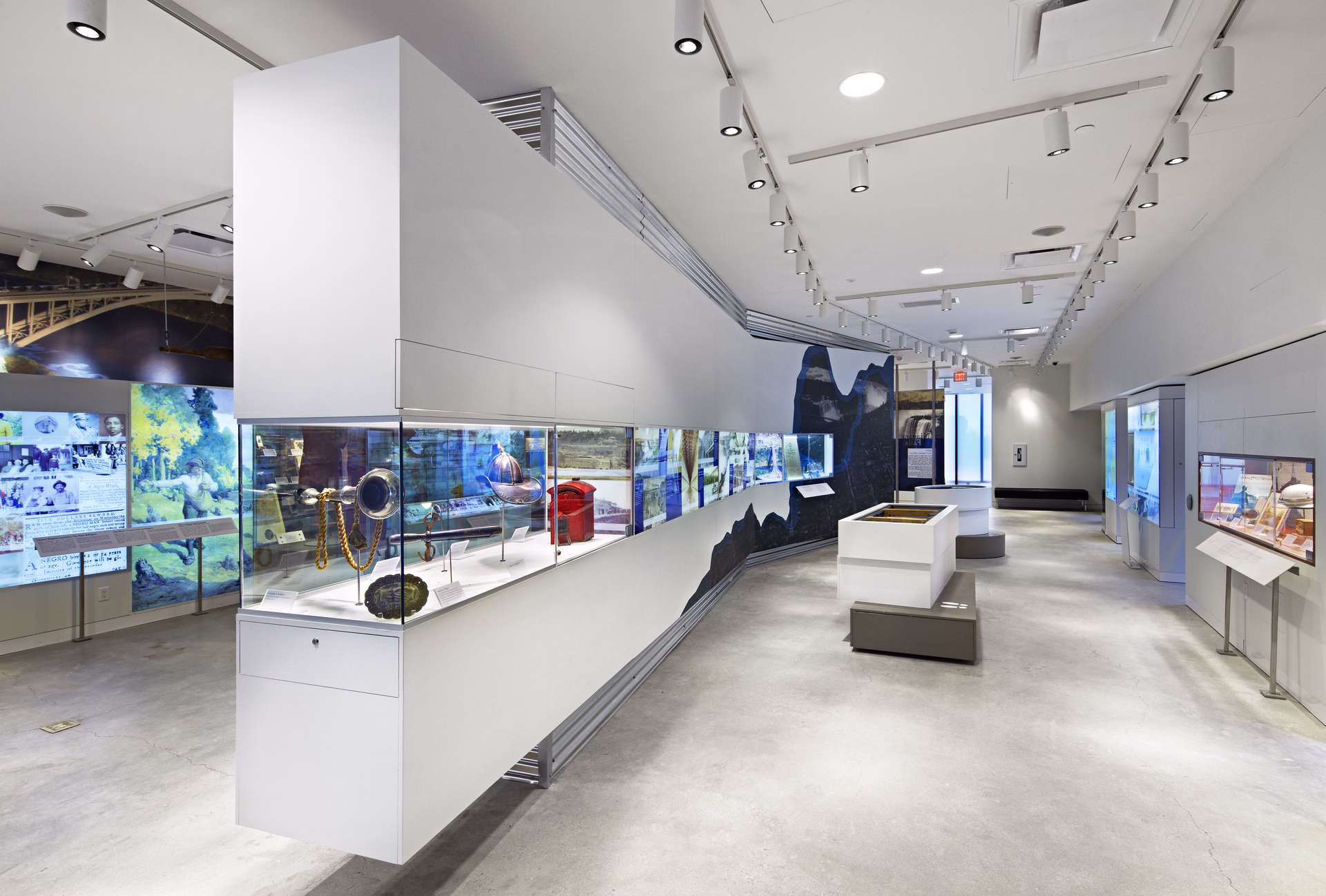 Modern museum gallery with long glass display cases featuring historical artifacts and illuminated panels on white walls.