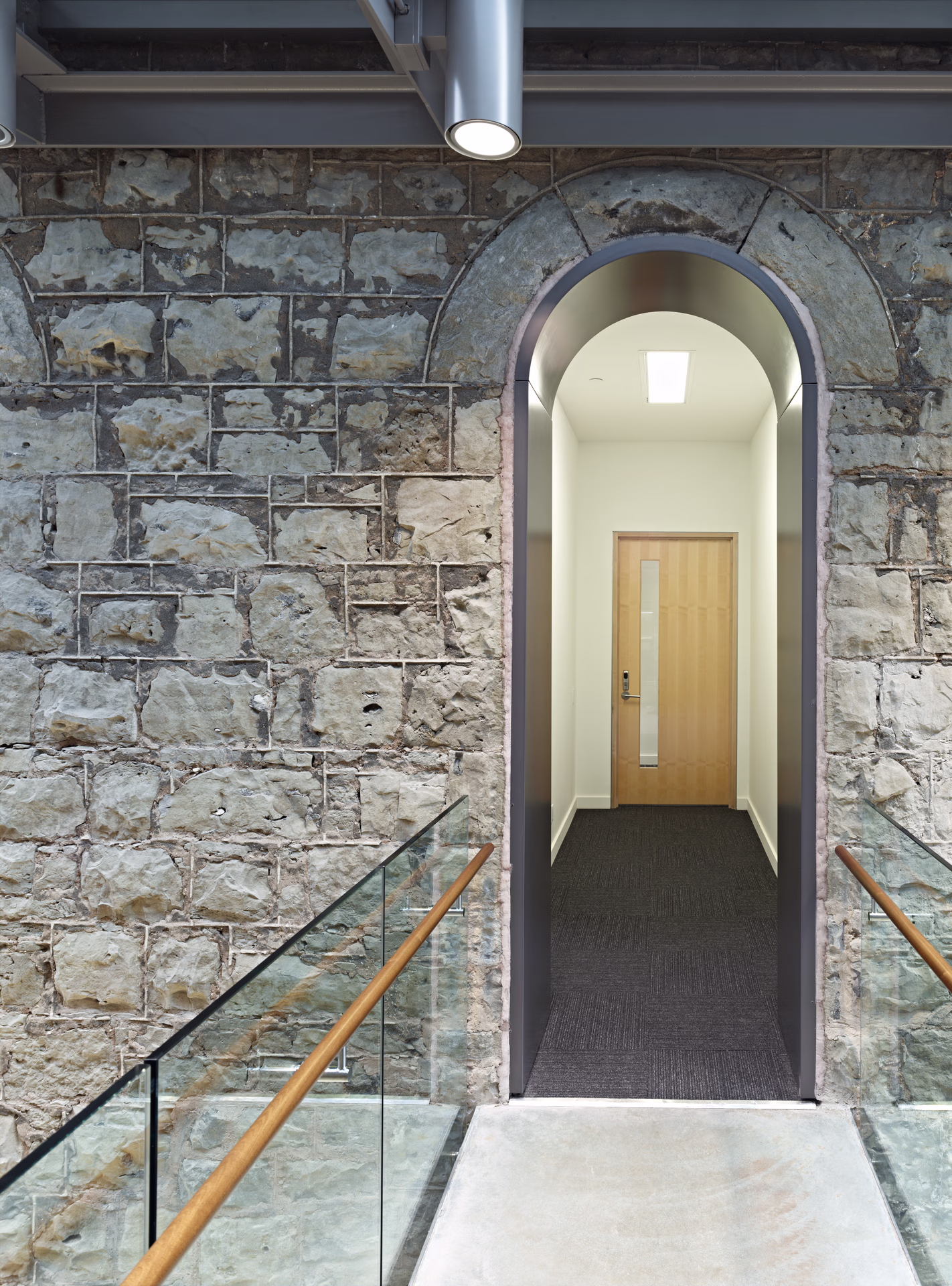 Stone wall with an arched doorway featuring a modern wooden door and a glass railing with wooden handrails on both sides.