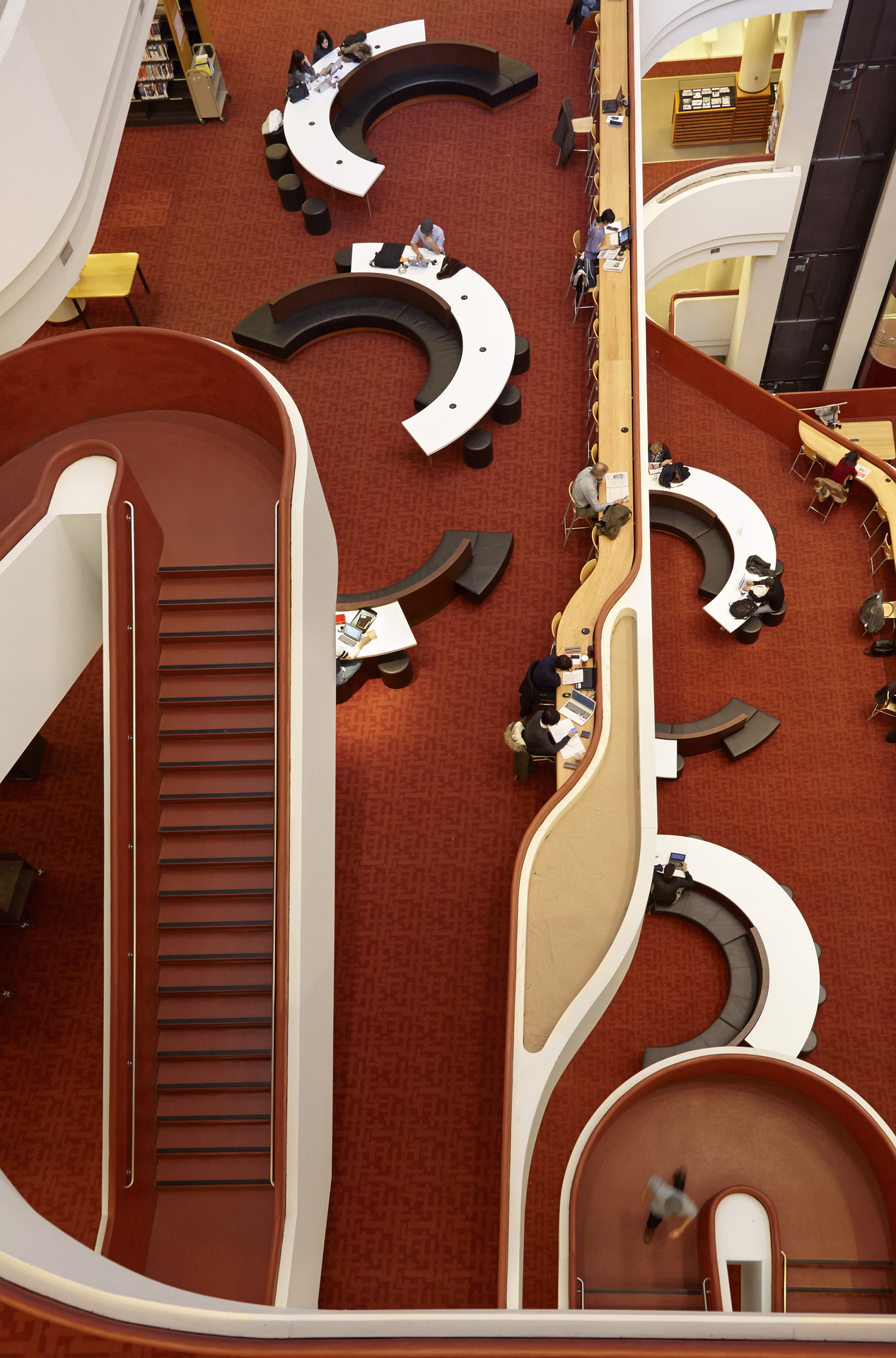 Overhead view of a modern library interior with curved white desks, red carpet, staircases, and people working on laptops.
