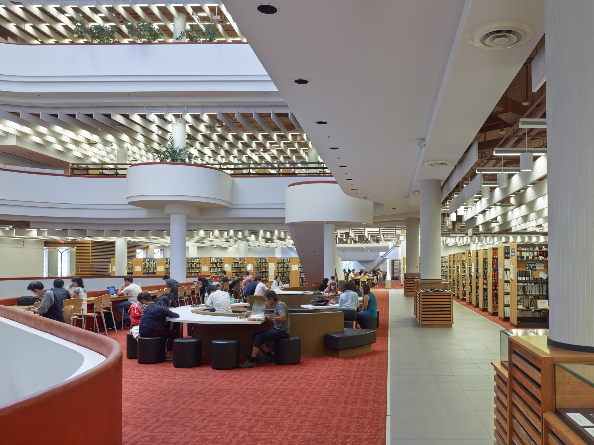 Spacious modern library interior with people studying at circular and rectangular tables under rows of white ceiling lights.