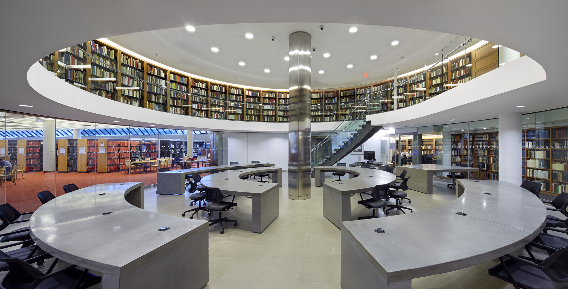 Modern library interior with circular concrete desks and black chairs under a mezzanine lined with bookshelves.