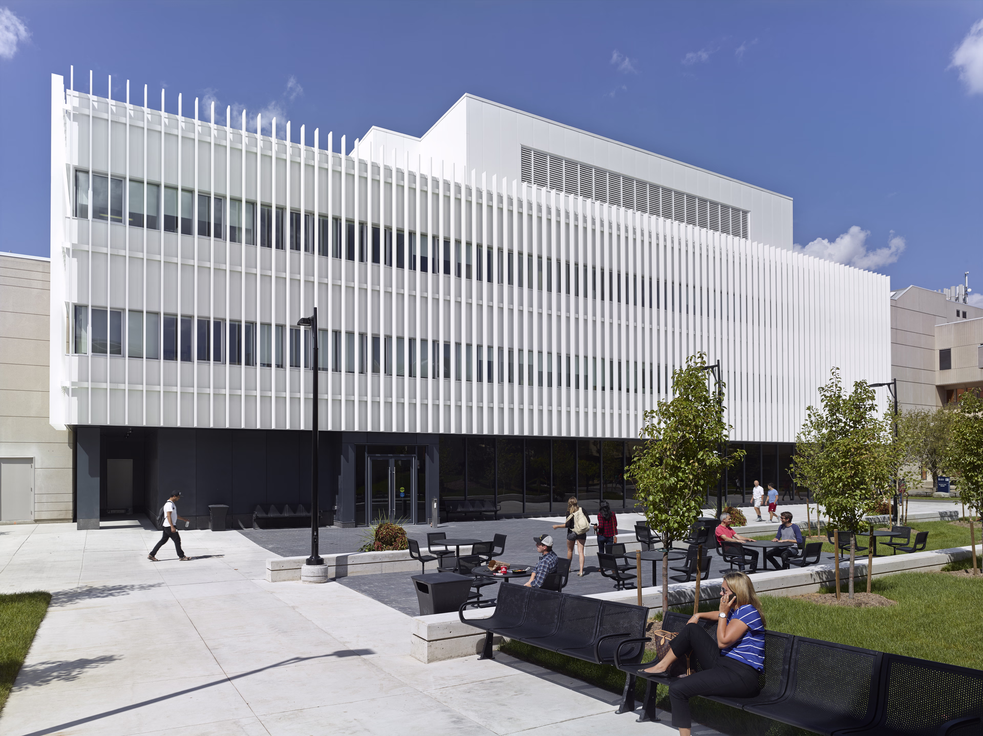 Modern white building with vertical slats, outdoor seating area with tables and chairs, and people walking and sitting under clear blue sky.