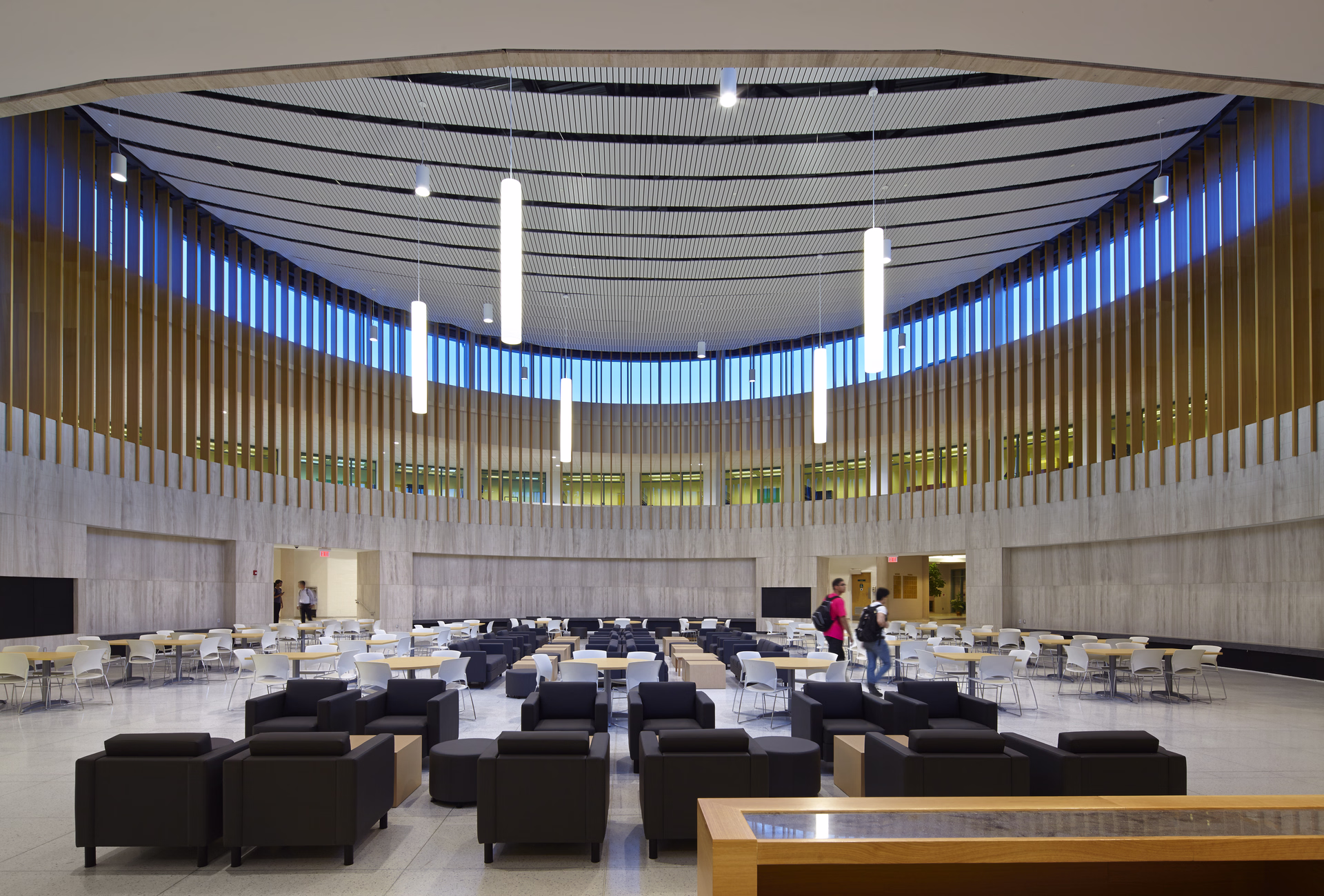 Spacious modern circular lounge area with black armchairs, wooden tables, white chairs, and tall vertical windows near ceiling.