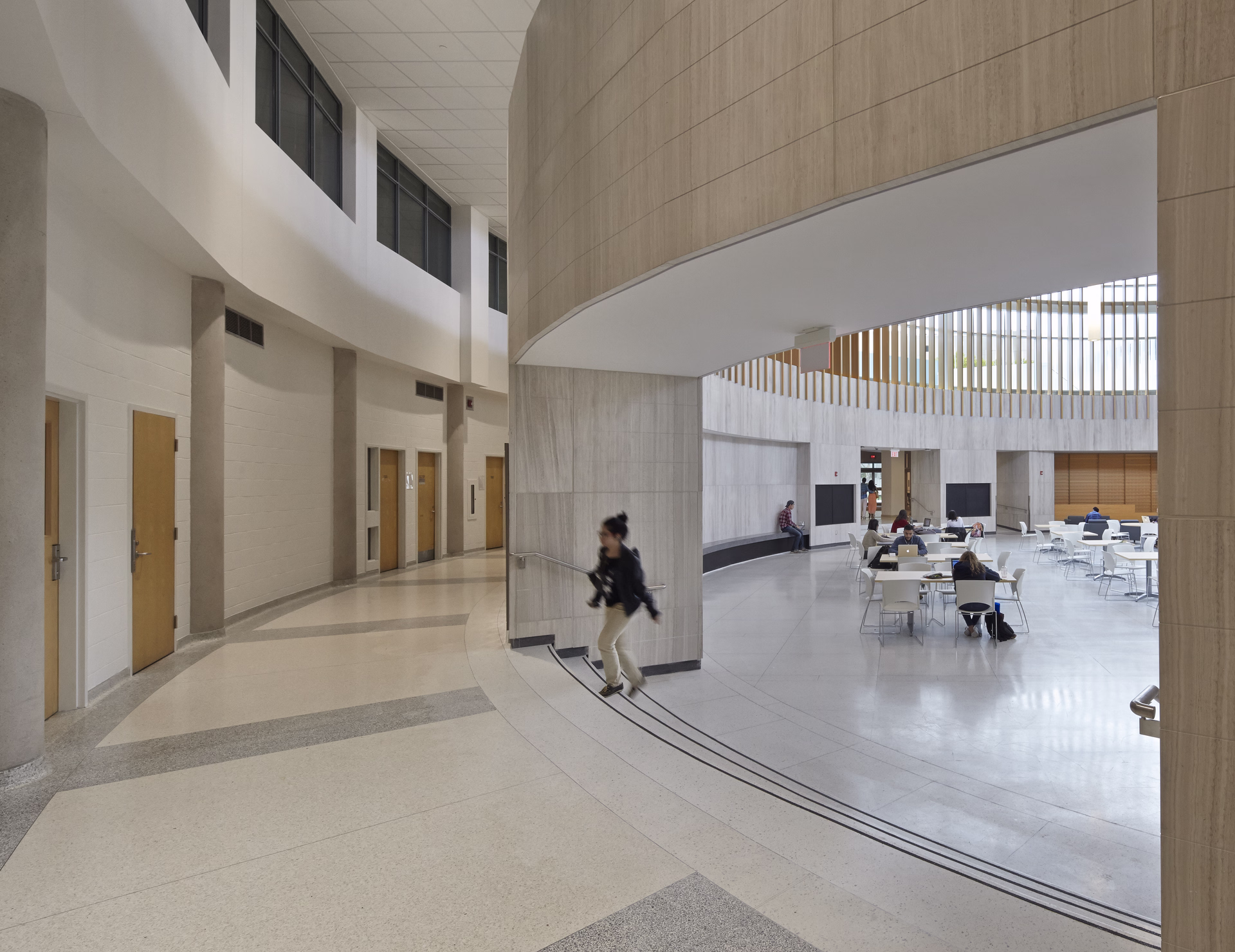 Modern indoor space with curved hallway, wooden doors, and an open atrium where people sit at tables working and socializing.