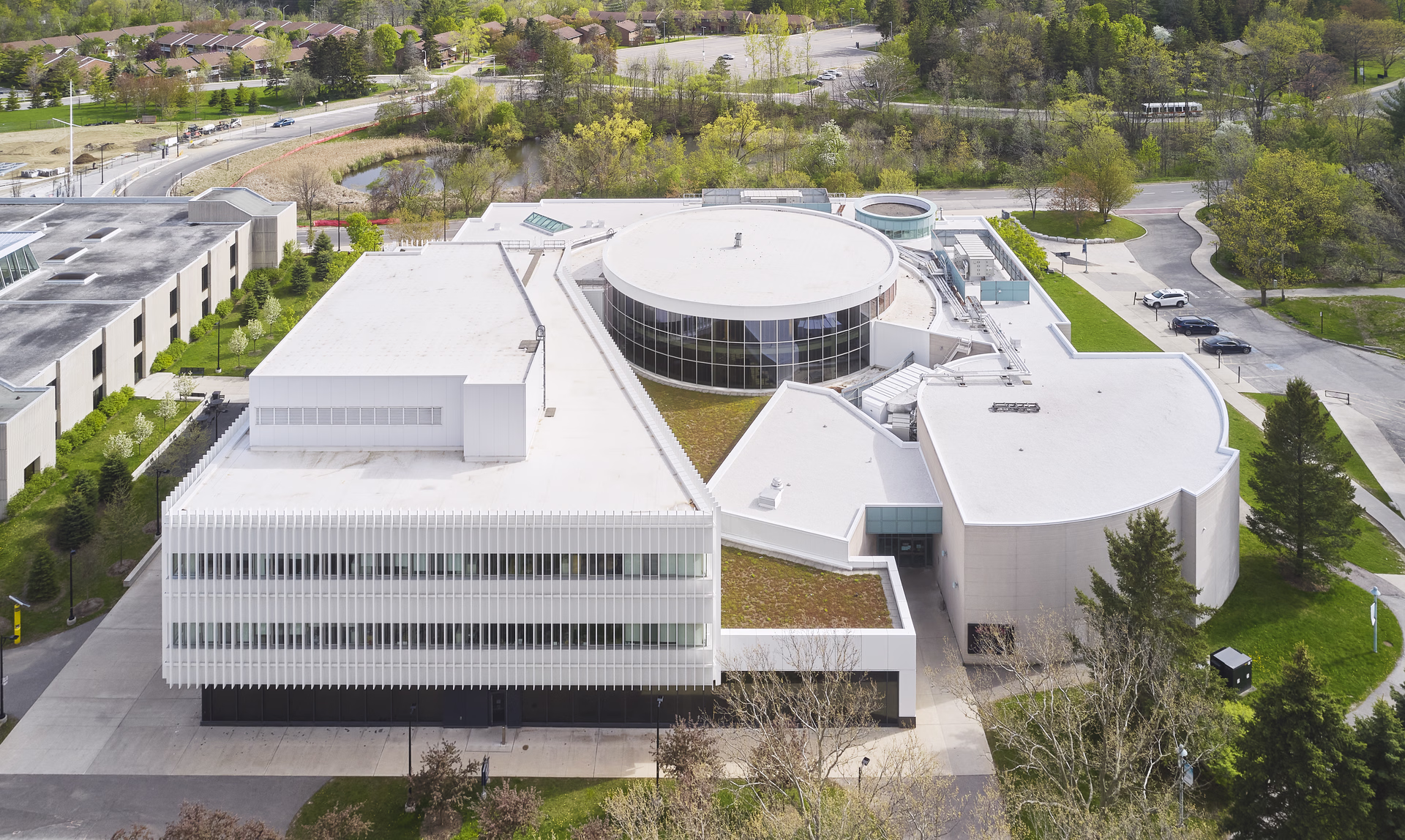 Aerial view of a modern white commercial building with circular and rectangular sections surrounded by trees and parking areas.