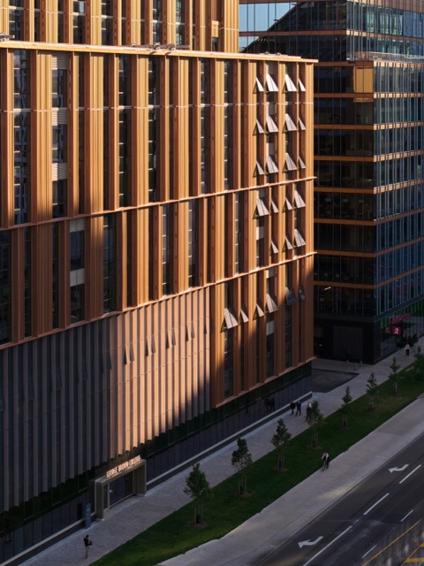 Modern office building with vertical wooden slats and reflective glass windows along a sidewalk and street.