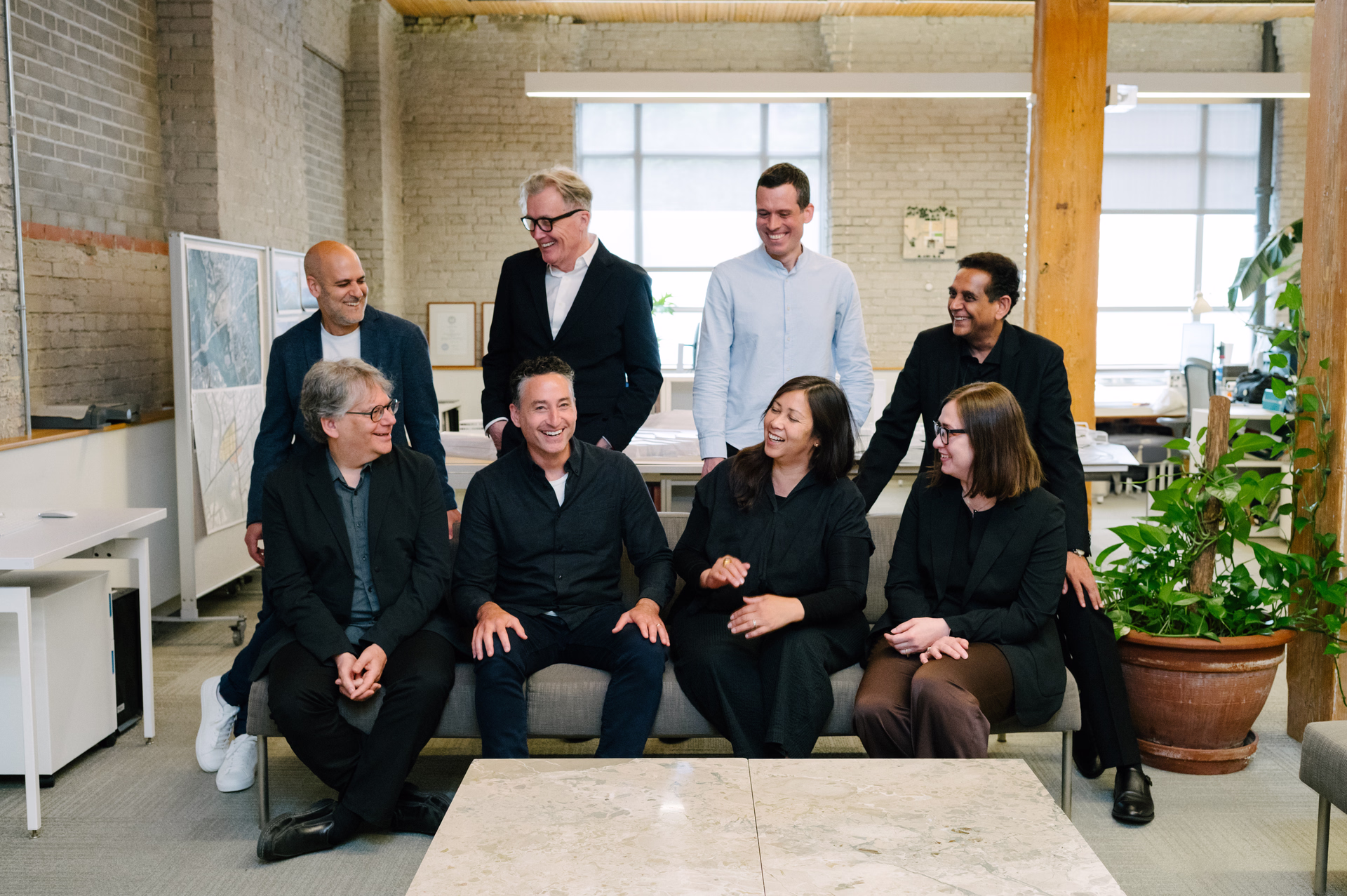 Group of eight casually dressed people sitting and standing together in a bright office space, smiling and chatting.