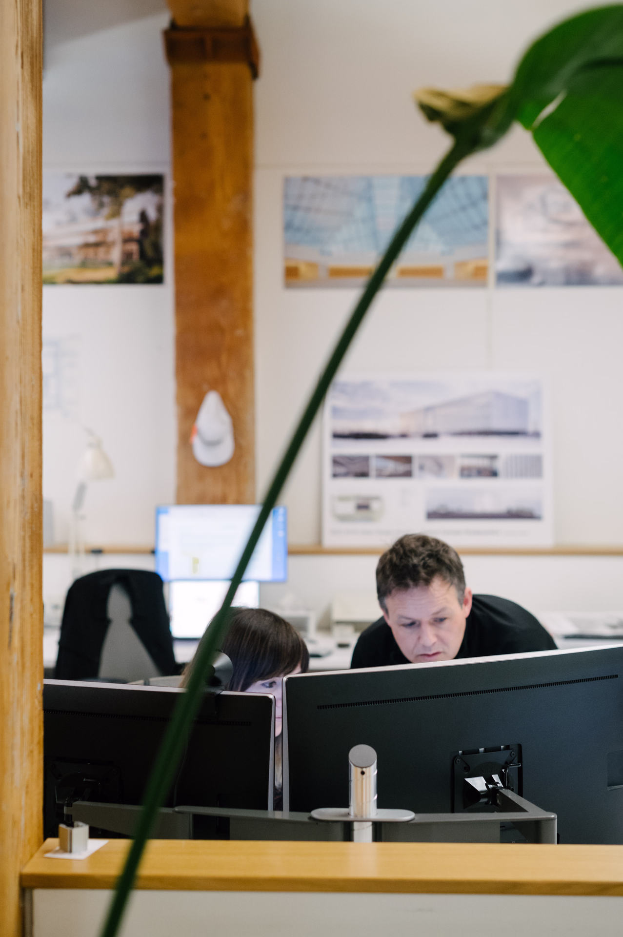 Two people collaborating while looking at computer screens in a bright office with architectural posters on the wall.
