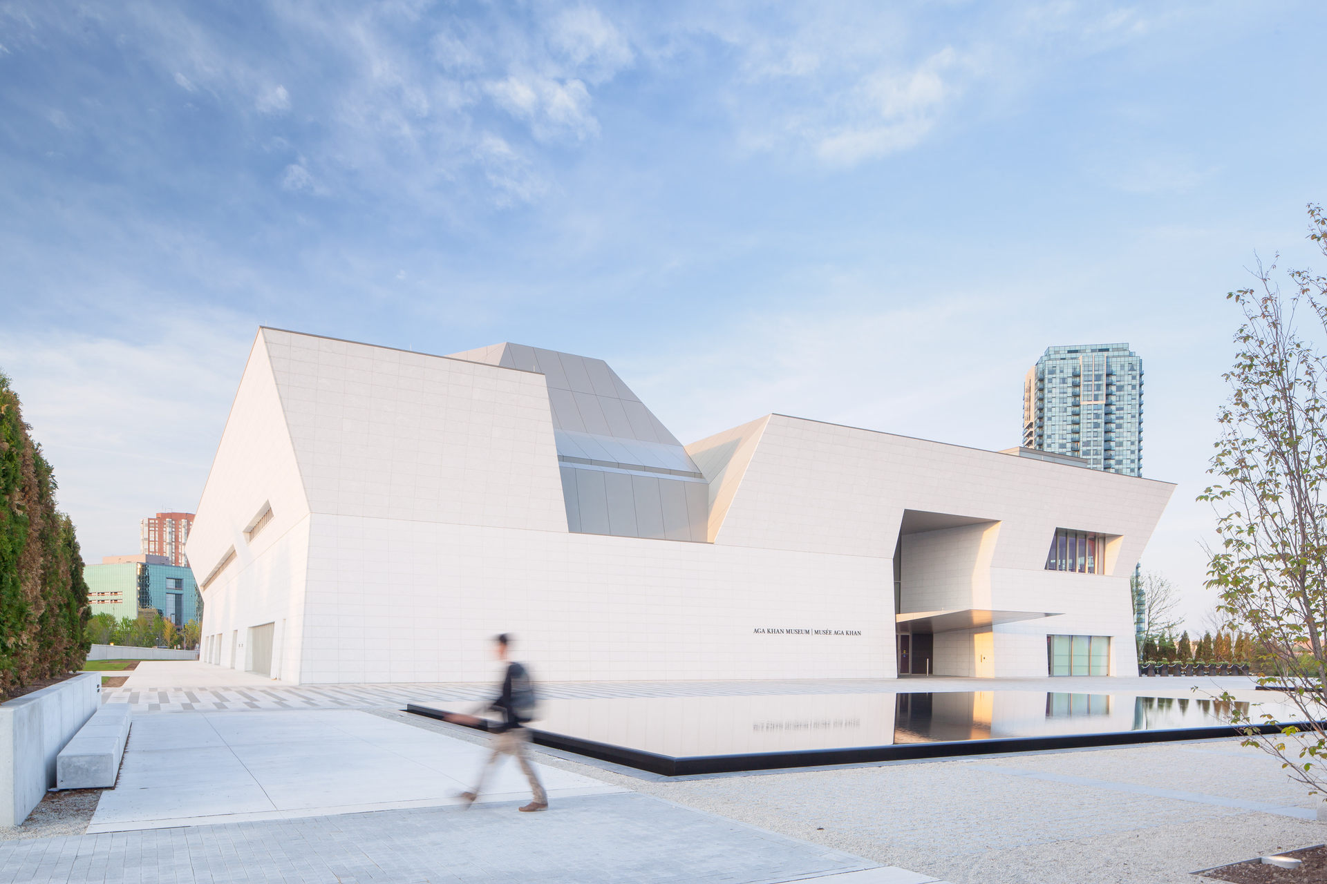 Modern white angular building of Aga Khan Museum with reflective water pool in front and a blurred person walking by.