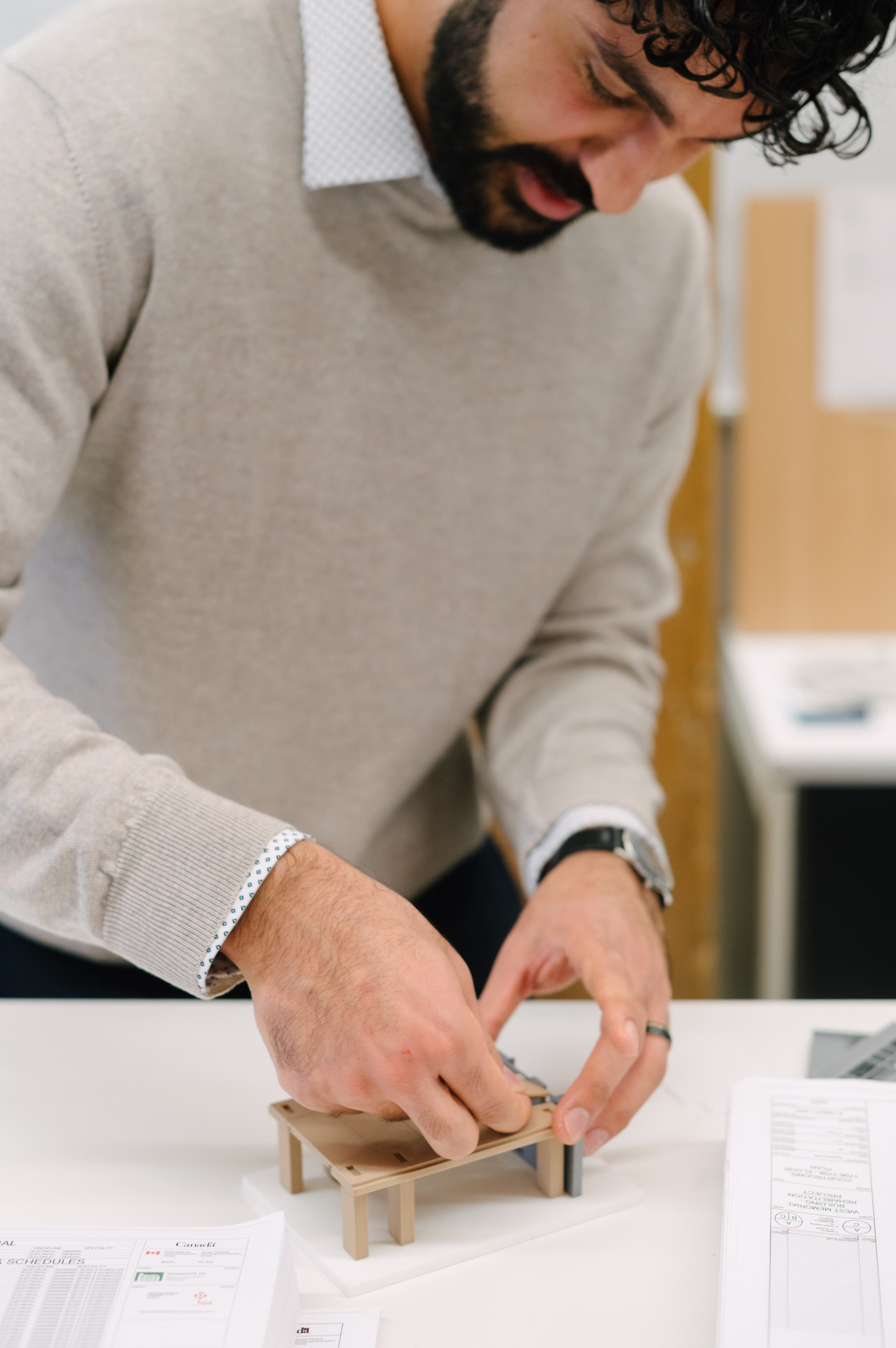 Man in a beige sweater adjusting a small wooden architectural model on a white desk with papers and documents nearby.