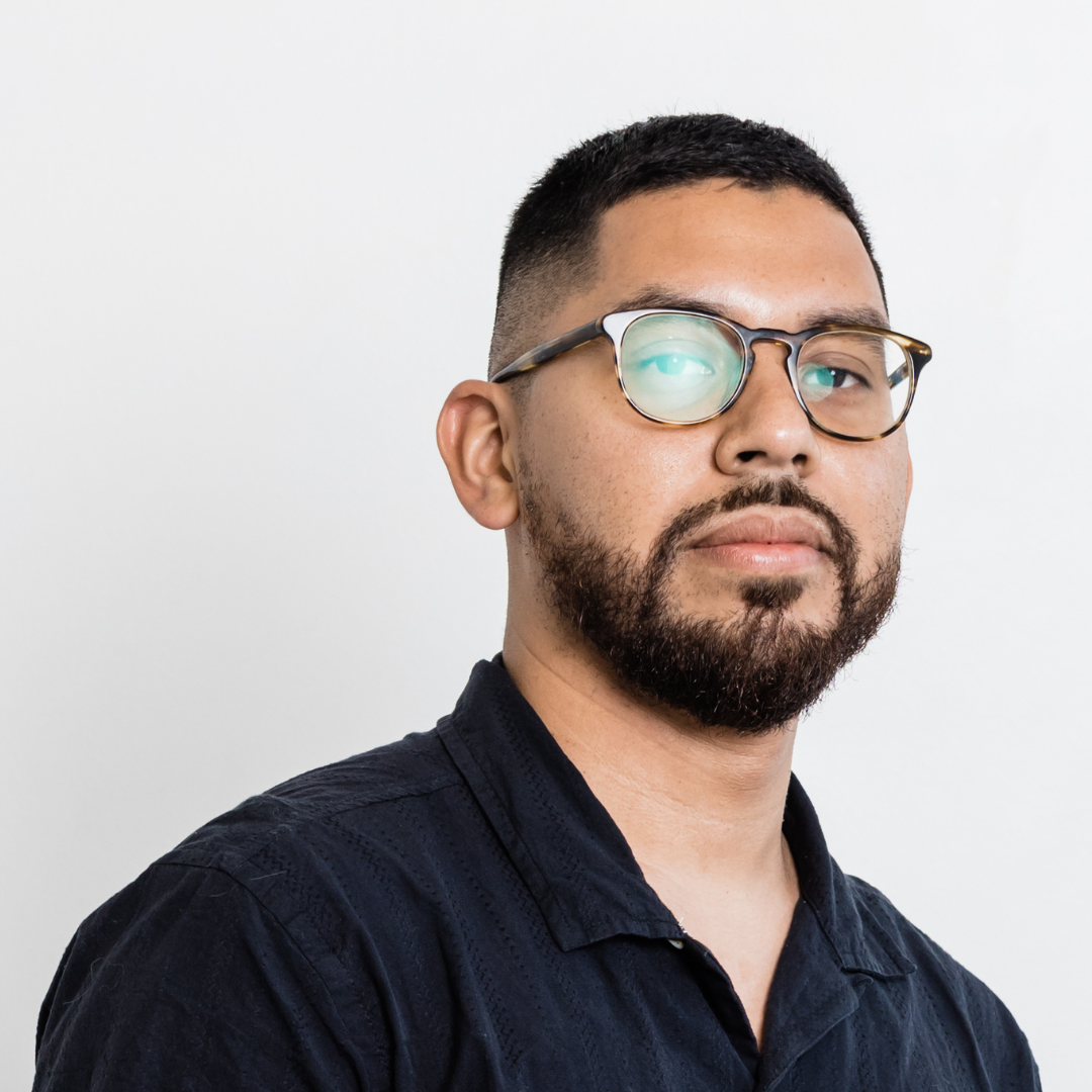 Portrait of a man with short black hair, beard, glasses, and wearing a dark shirt against a plain white background.