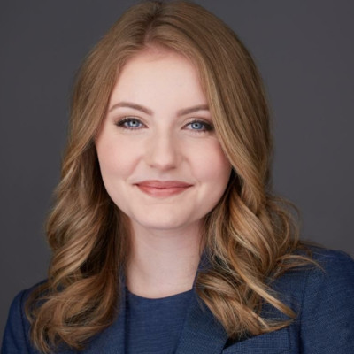 Smiling woman with light brown wavy hair wearing a blue blazer against a gray background.