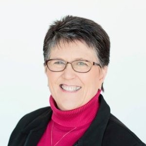 Smiling woman with short dark hair, glasses, wearing a pink turtleneck and black jacket against a white background.